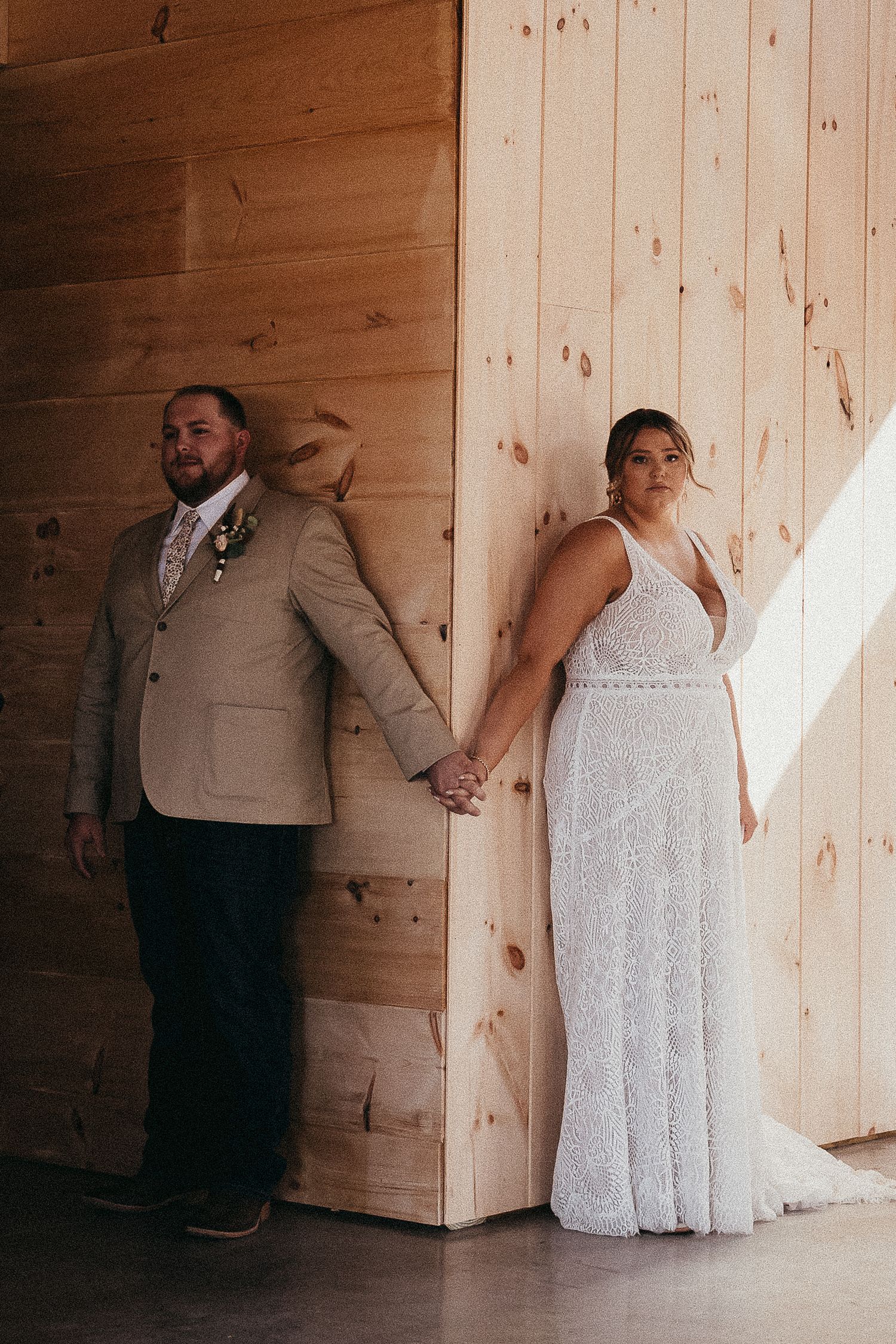 Wedding couple holding hands against rustic wood wall, bride in lace gown and groom in tan suit.
