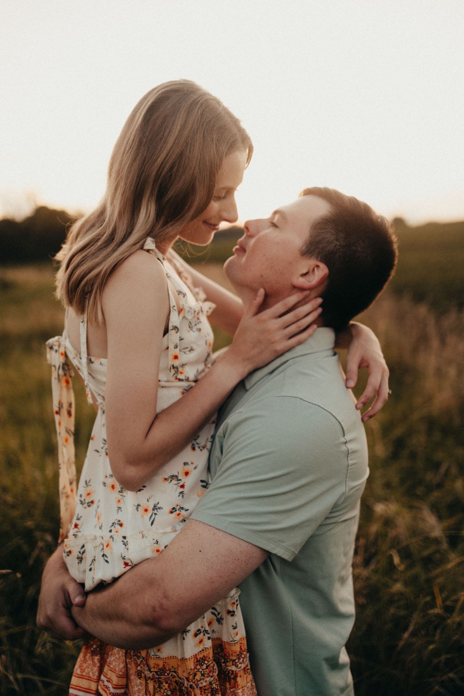 Couple nearly kissing at sunset in a meadow, woman in floral dress held by man in green shirt.