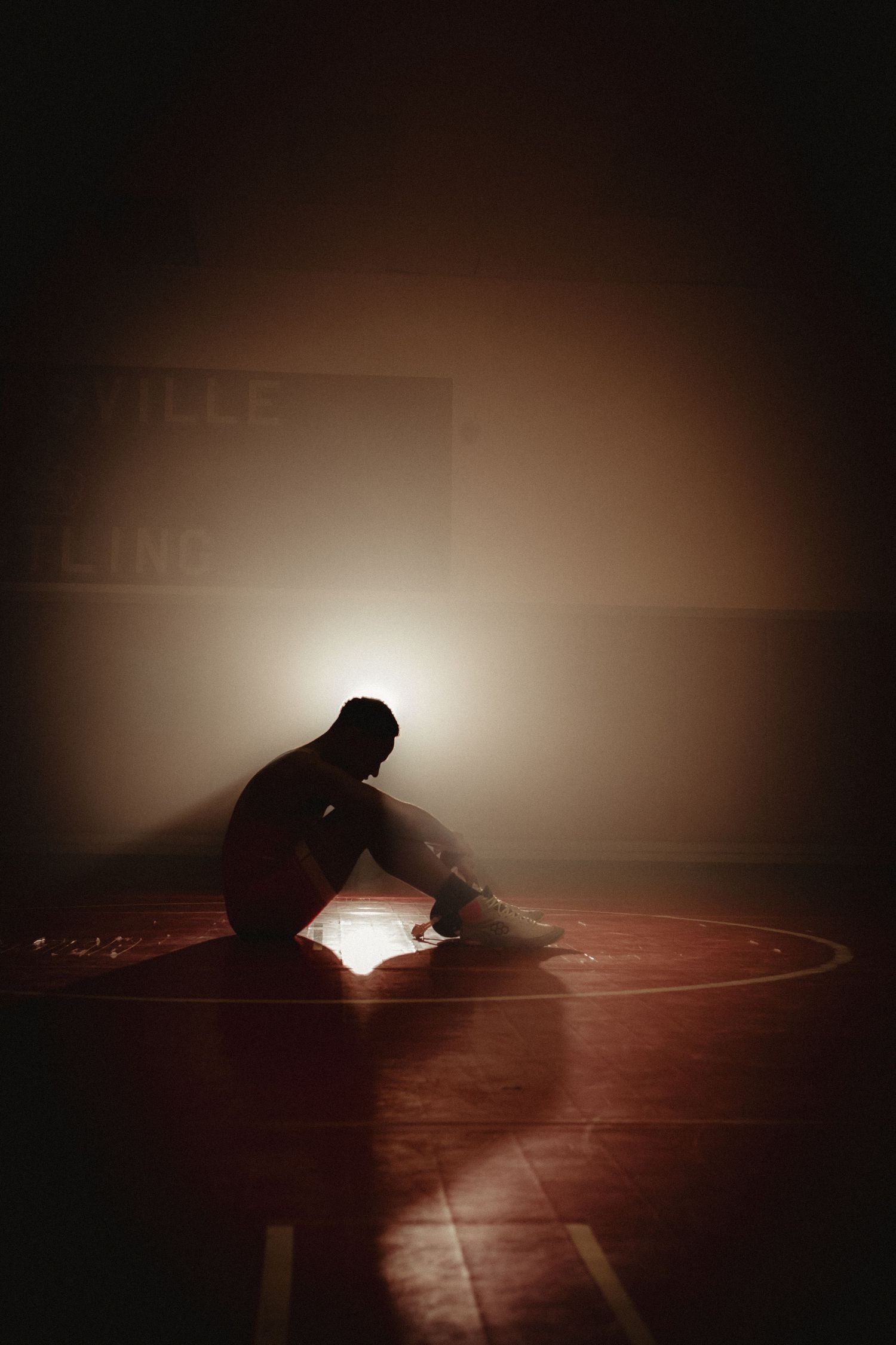 Silhouette of a wrestler sitting alone on a mat under a single spotlight in a dark gymnasium.