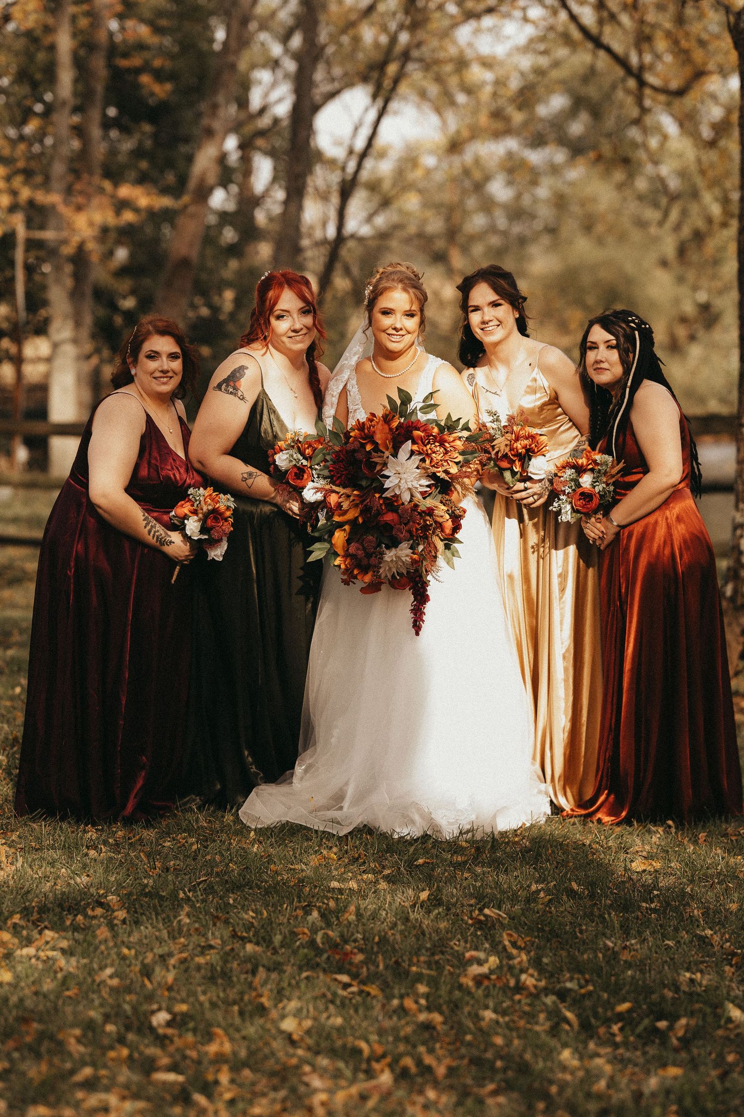 Bride in white gown poses with four bridesmaids in mismatched jewel-tone velvet dresses holding fall floral bouquets outdoors.
