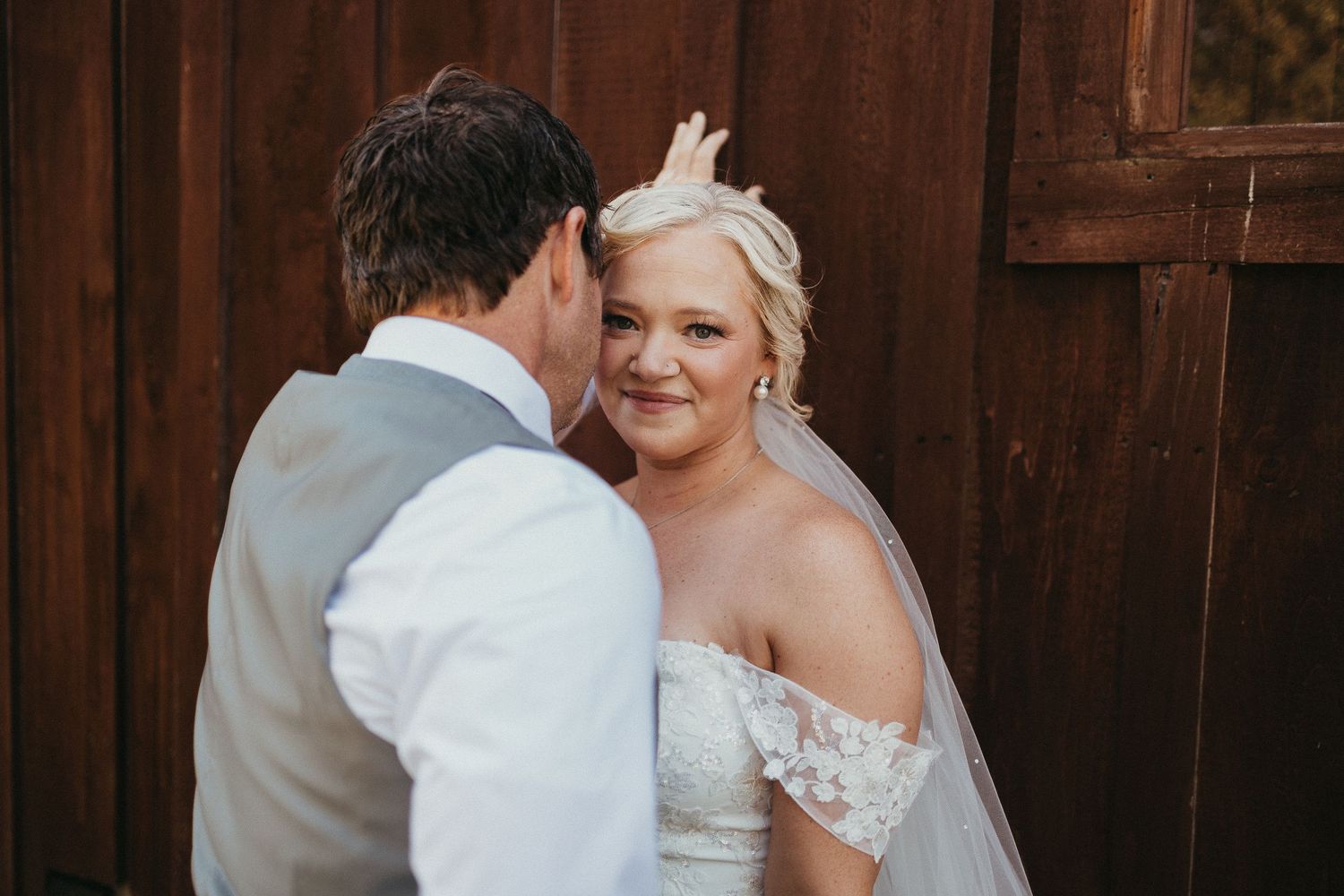Bride in off-shoulder lace gown with veil smiling at groom in gray vest outside rustic wooden barn wall.