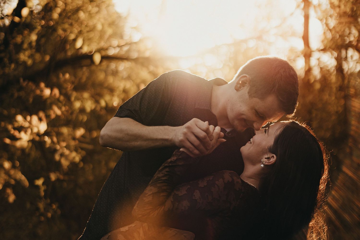 Romantic couple holding hands and leaning in for a kiss outdoors, glowing in warm golden sunset light among autumn trees.