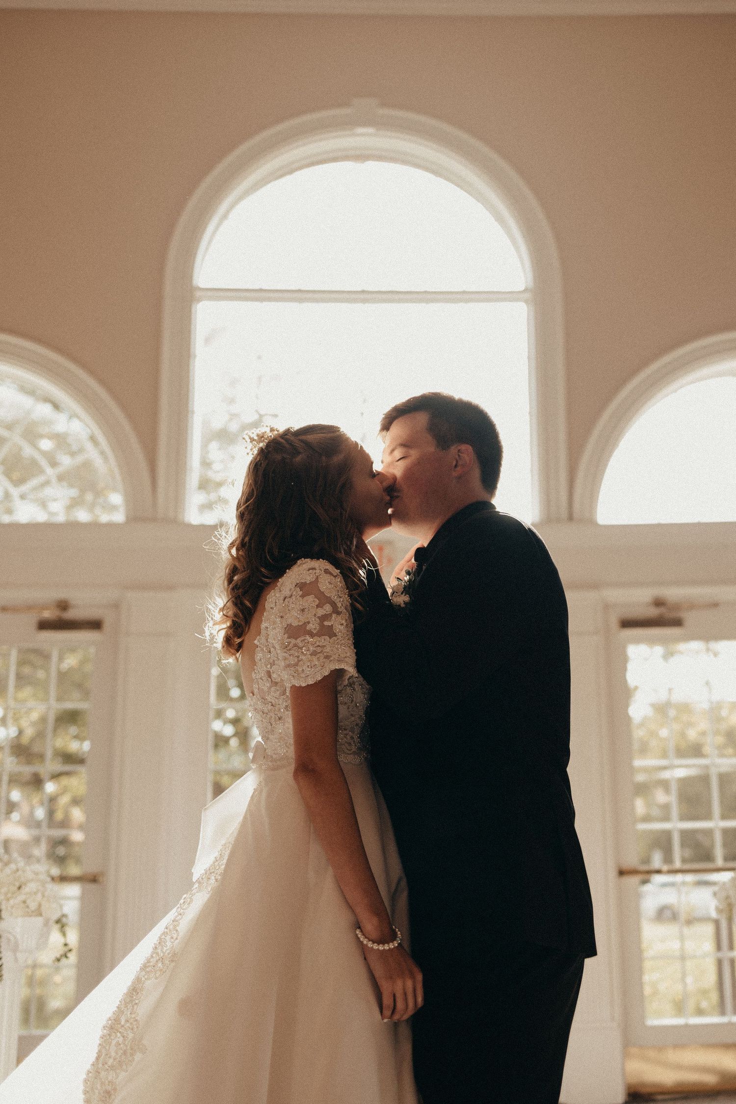 Bride in lace gown and groom share first kiss inside elegant venue with arched windows and natural light.