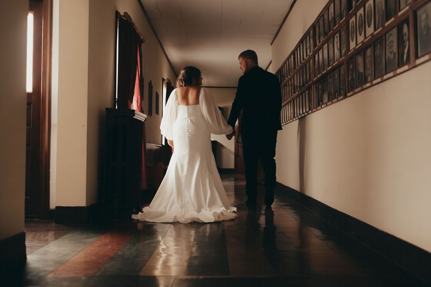 Bride and groom walking hand in hand down hallway lined with framed portraits.