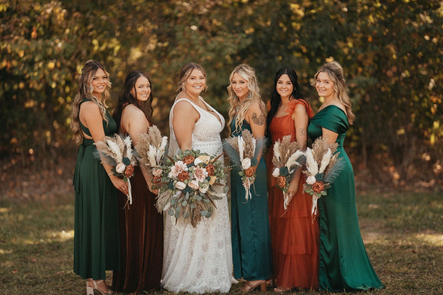 Bride with bridesmaids in emerald, burgundy, and rust gowns holding bouquets in fall foliage setting.