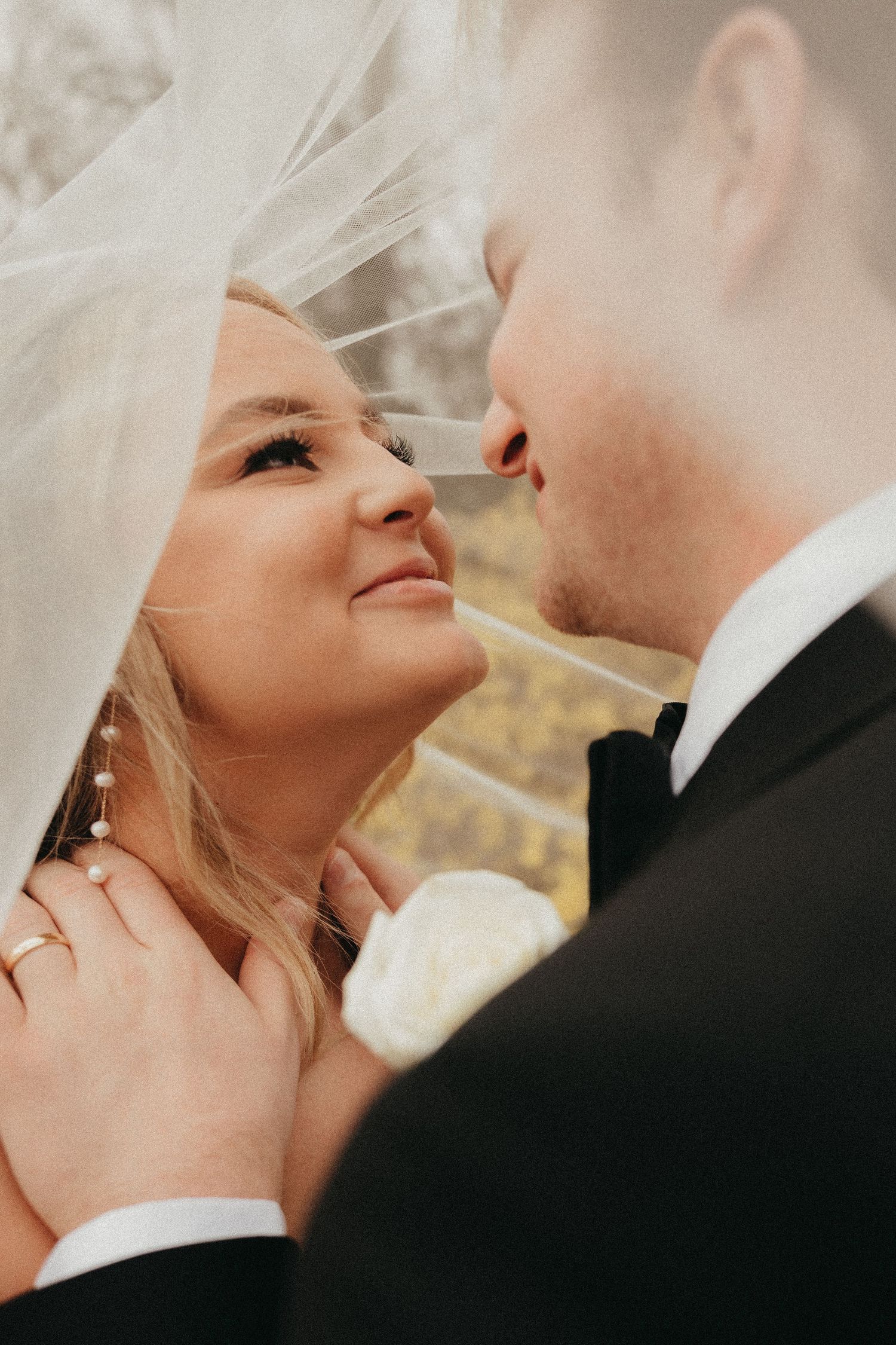 Bride gazes lovingly at groom under flowing white veil wearing pearl earrings and wedding ring.