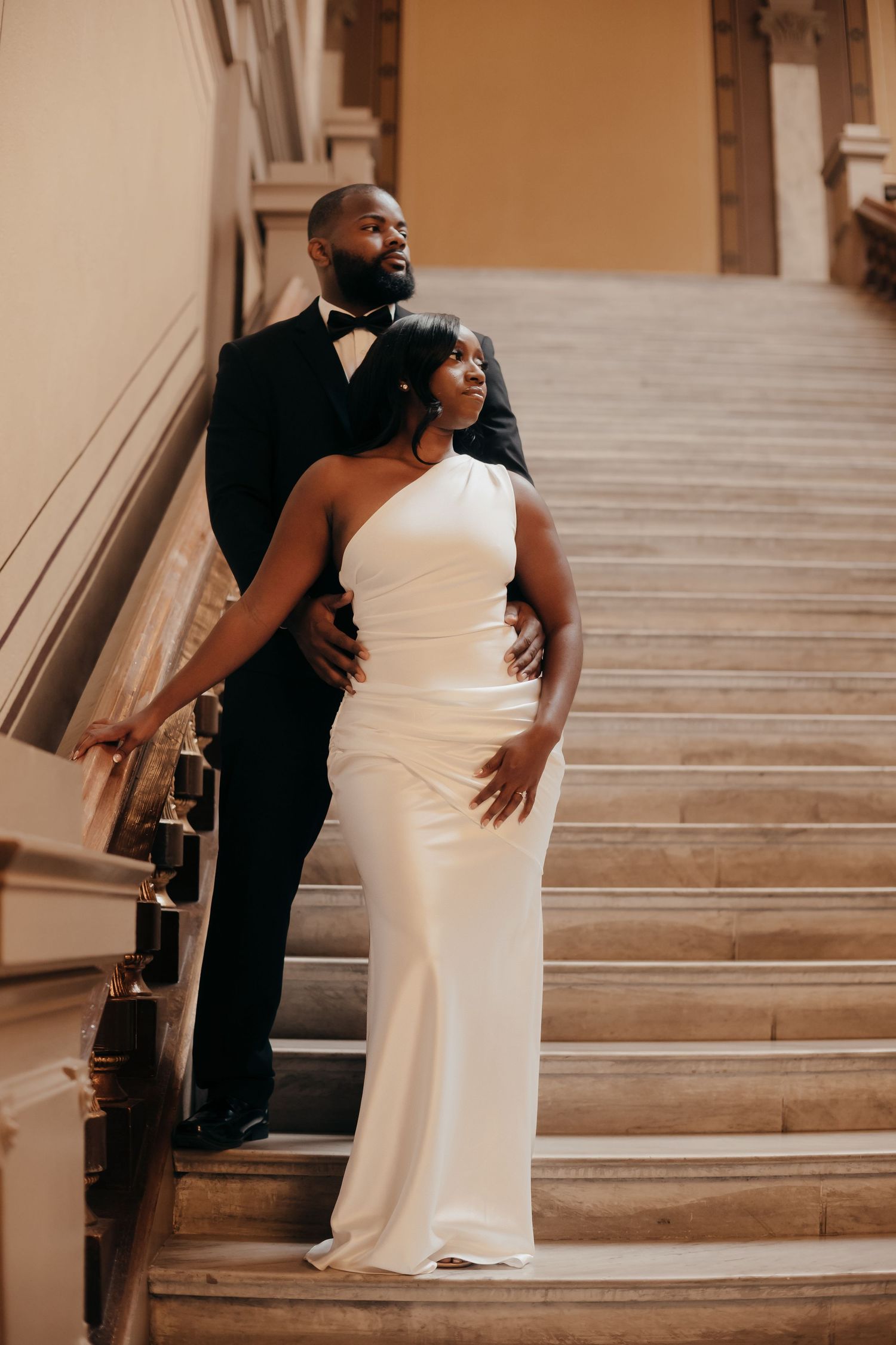 Couple in formal wedding attire pose on a grand stone staircase, the bride in a white one-shoulder gown and the groom in a black tuxedo.