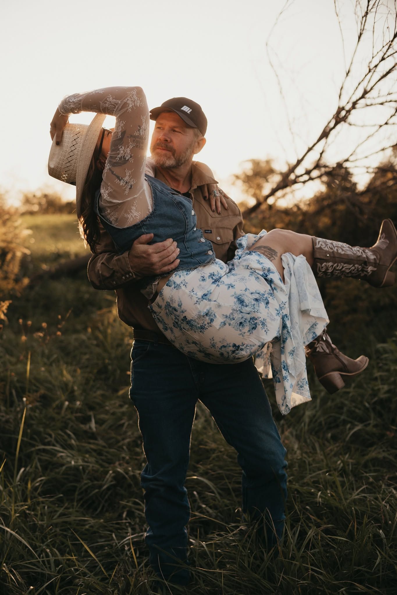 A man carries a woman in his arms in a sunlit field, both dressed in rustic western attire at golden hour.