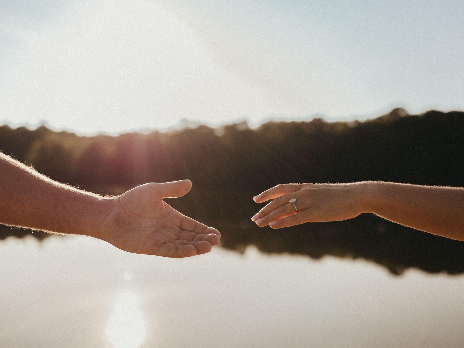 Two hands reaching toward each other against a sunlit lake backdrop, one adorned with a diamond engagement ring.
