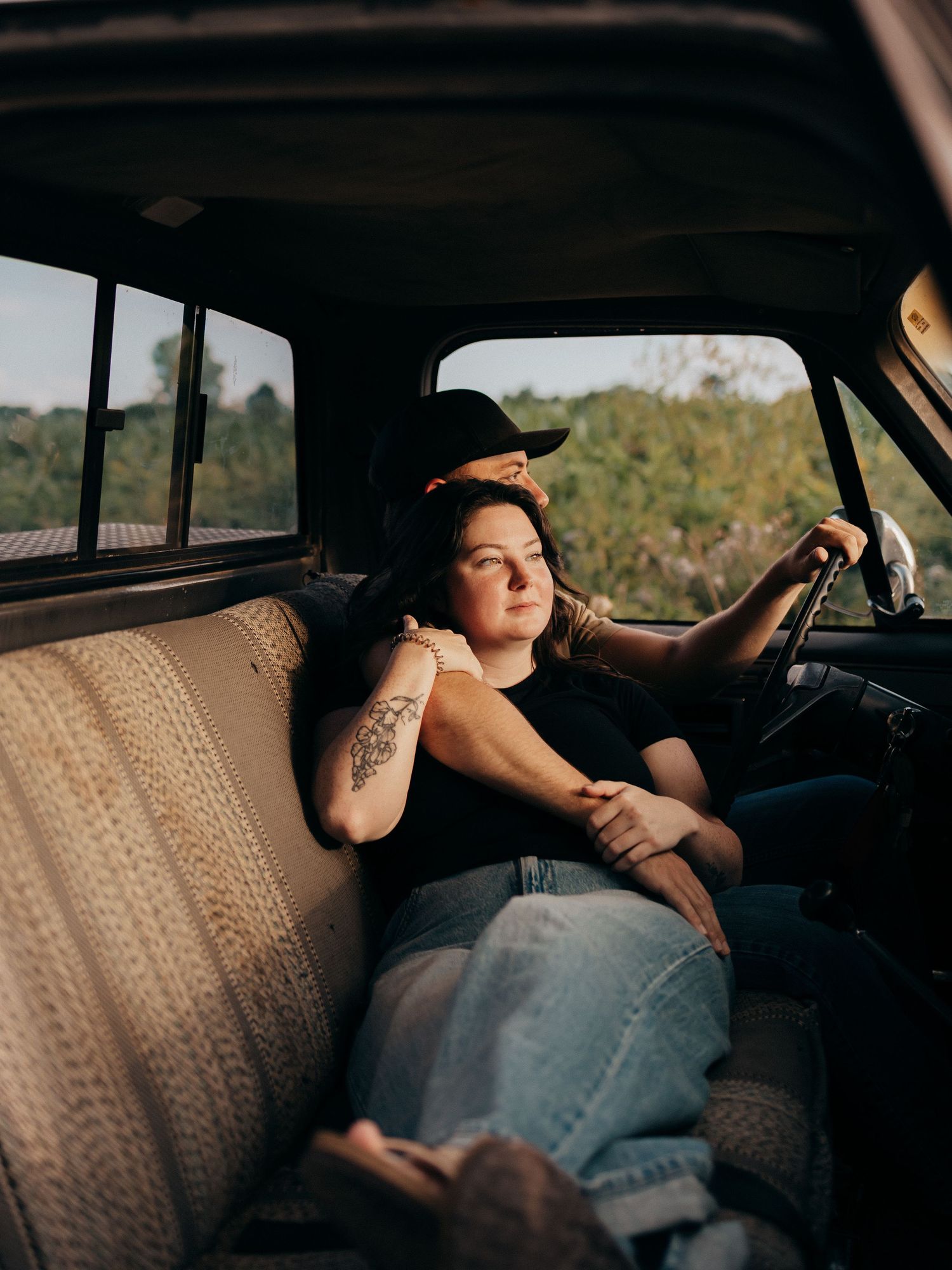 Woman with tattoos and black cap sits relaxed in vintage truck cab, golden hour light streaming through windows.