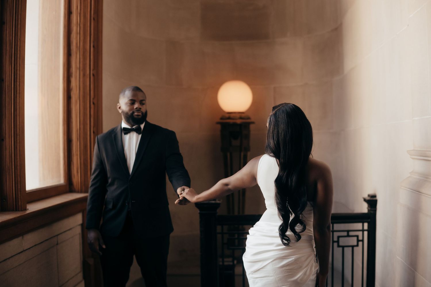 A groom in a black tuxedo and bow tie holds hands with a bride in a white dress with long dark hair inside an elegant, softly lit hall.