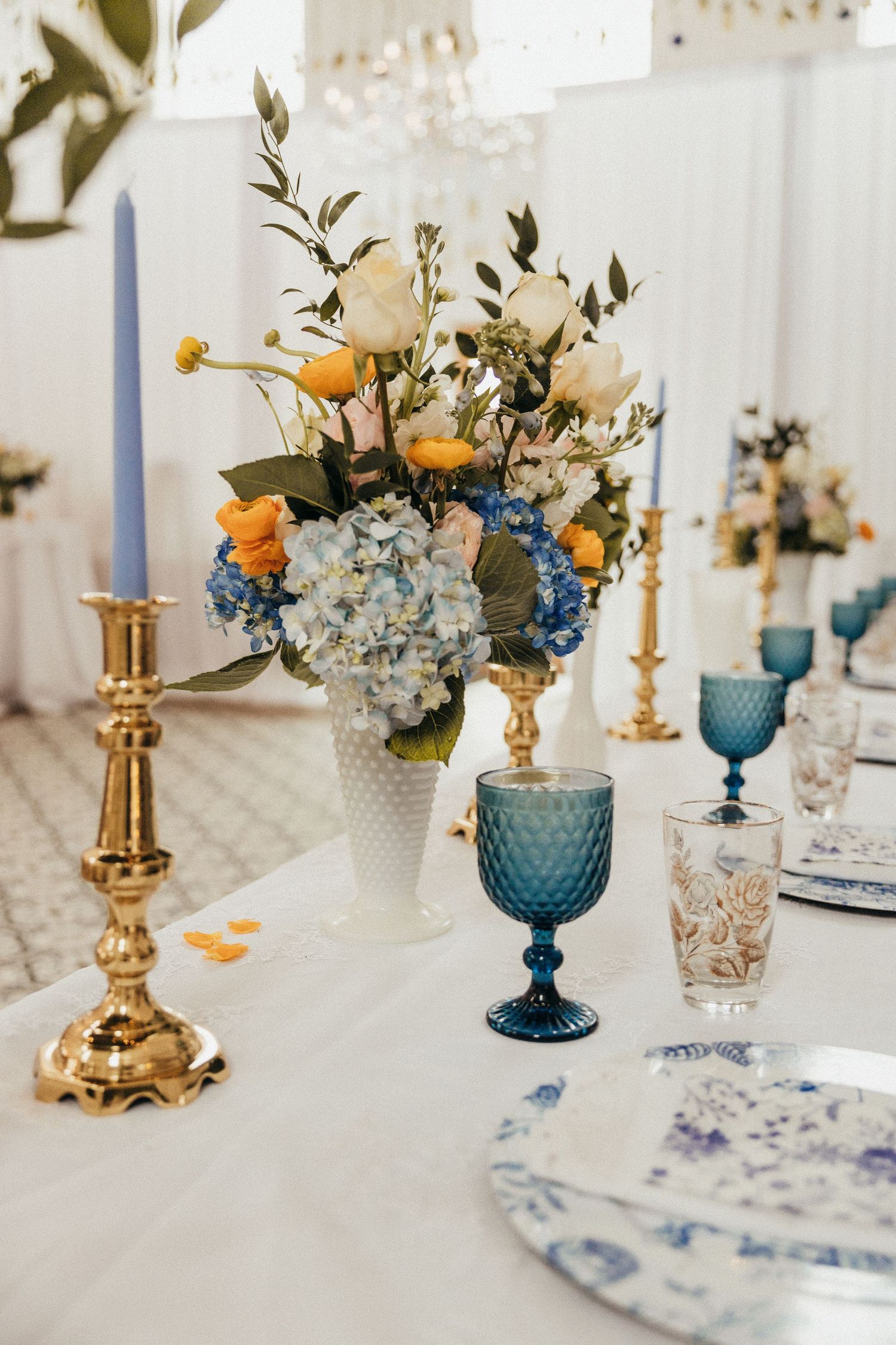 Elegant wedding tablescape with blue hydrangeas, orange roses, gold candlesticks, and blue goblets.