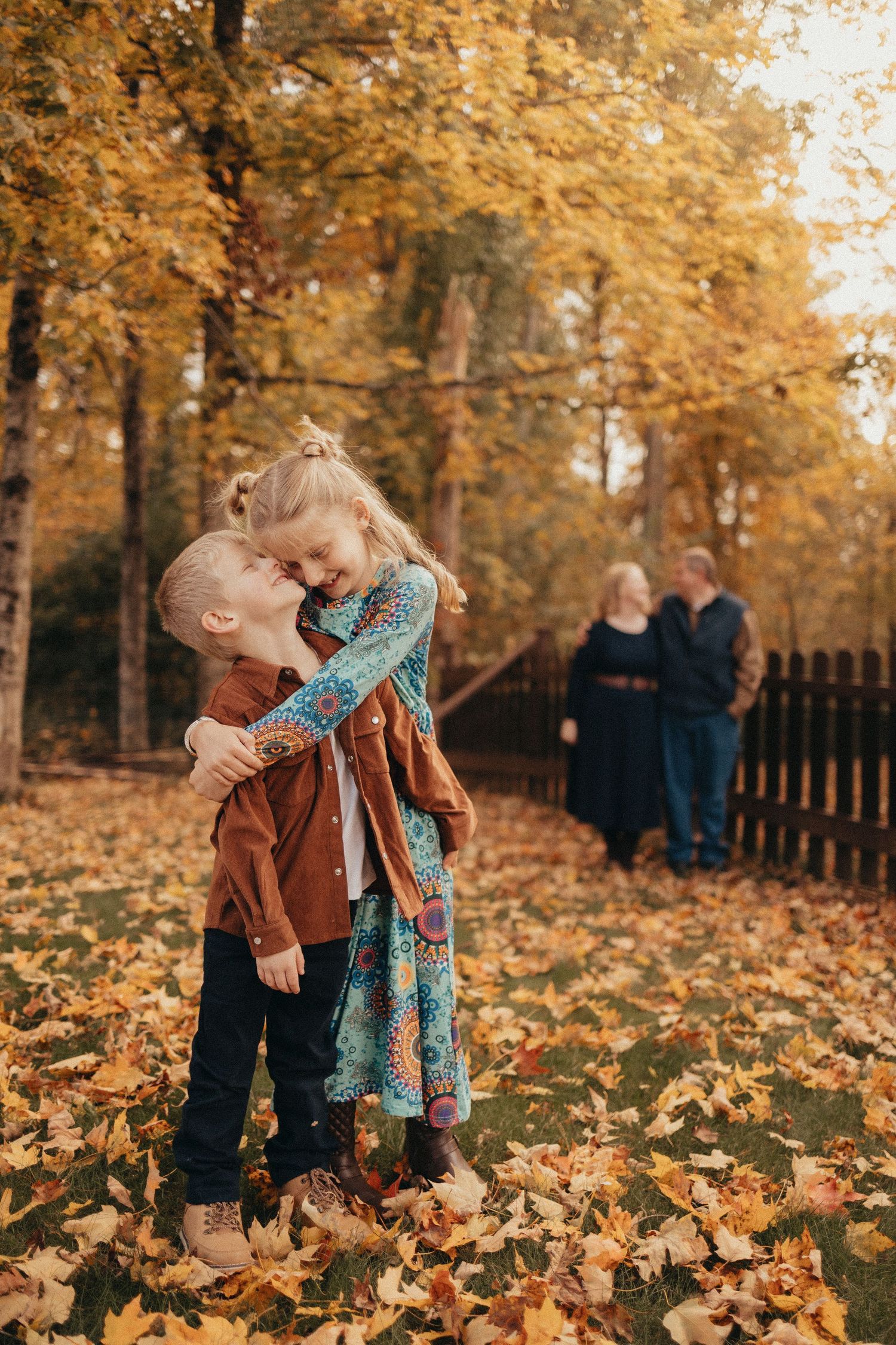 Two young children share a sweet embrace among fallen autumn leaves with grandparents standing in the background.