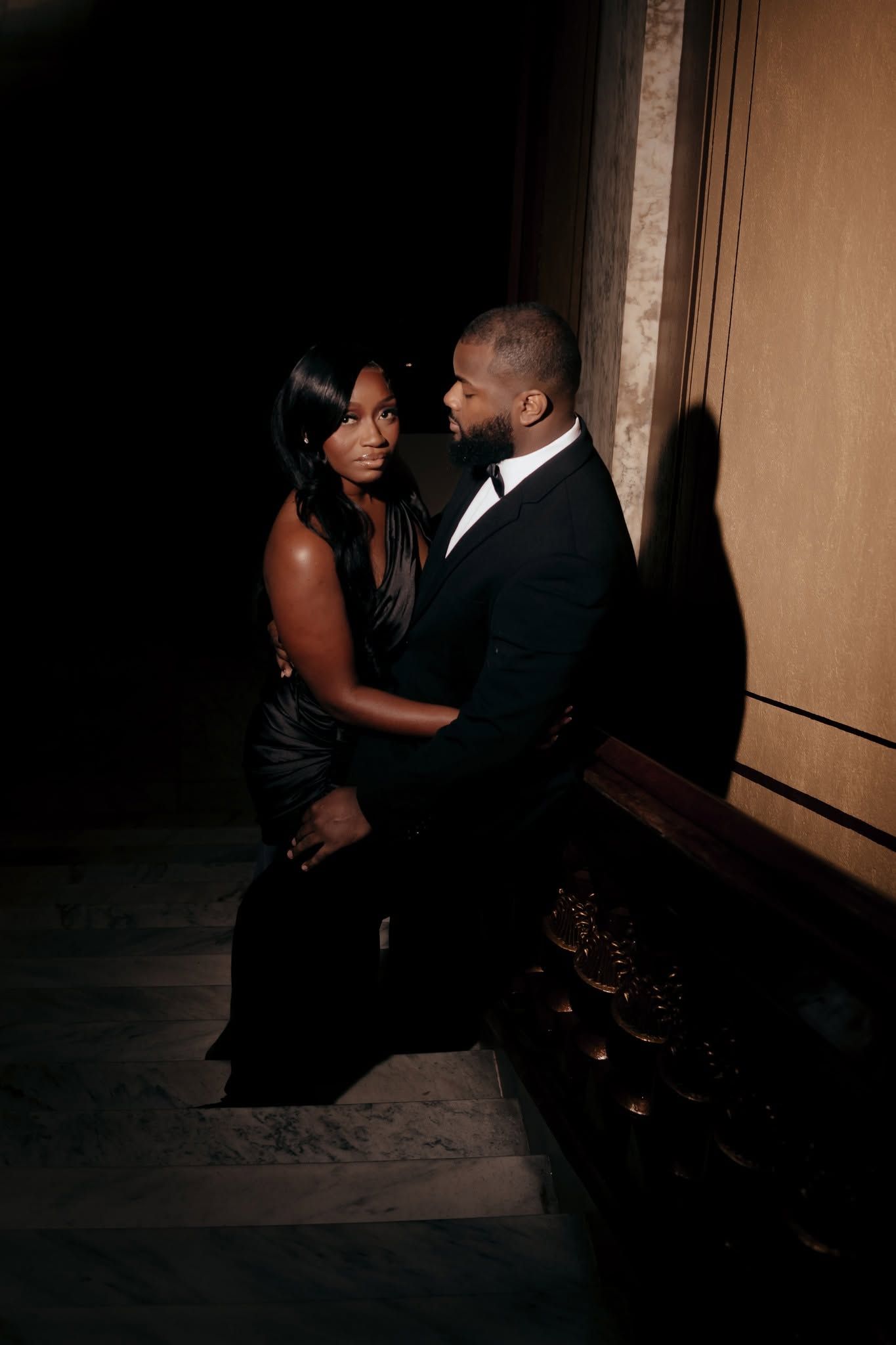 Elegant couple in formal black attire sharing an intimate moment on dimly lit stairs in a sophisticated venue.