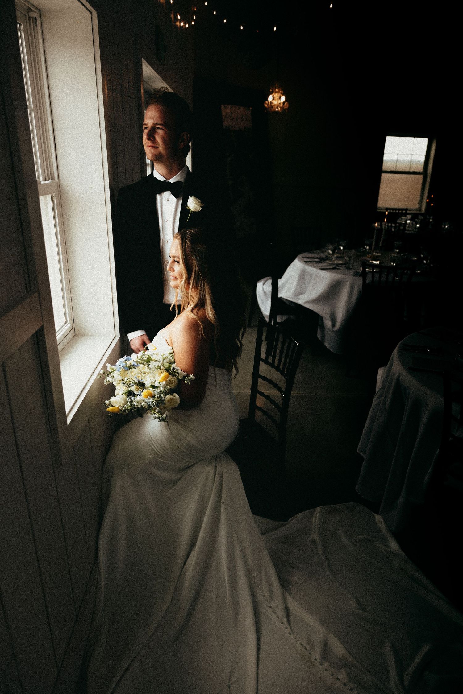 Bride and groom share a romantic moment by a window, bride holding white bouquet in elegant wedding gown.