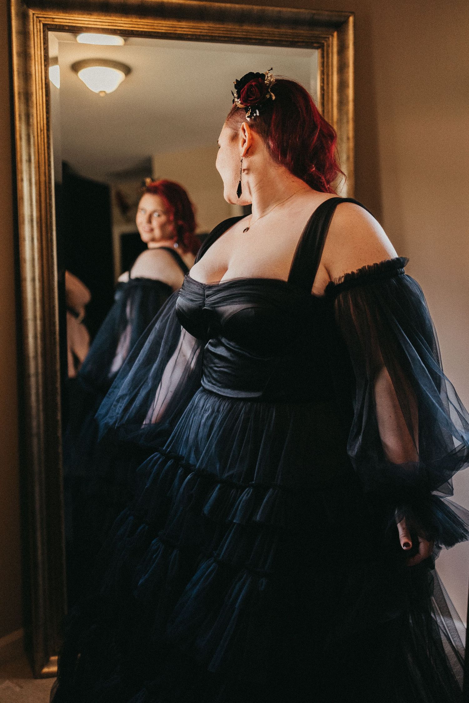 Woman in a dramatic black off-shoulder tulle ball gown with puff sleeves admires her reflection in a gold-framed mirror.