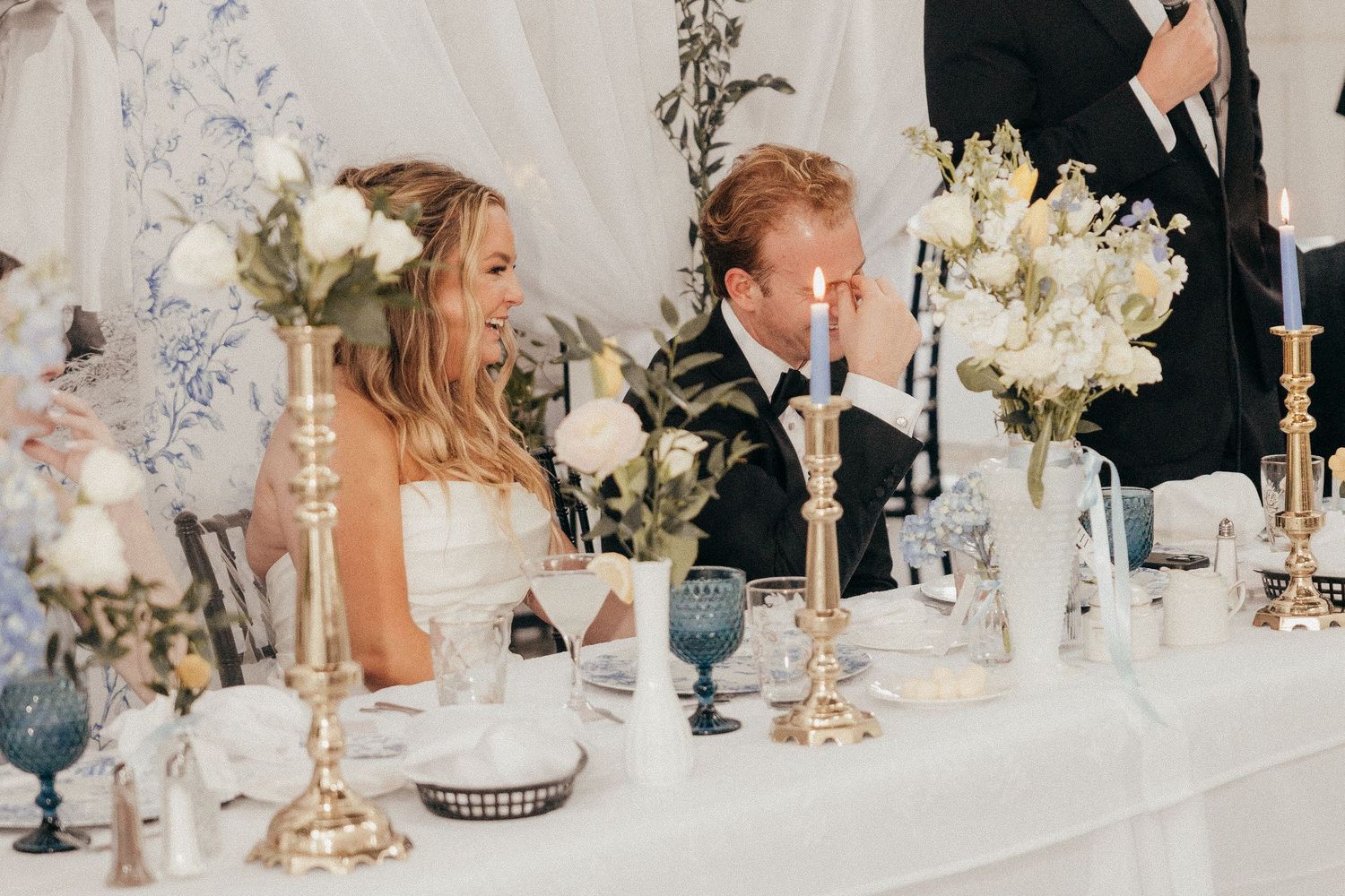 Bride and groom smile at sweetheart table adorned with white florals and gold candlesticks.