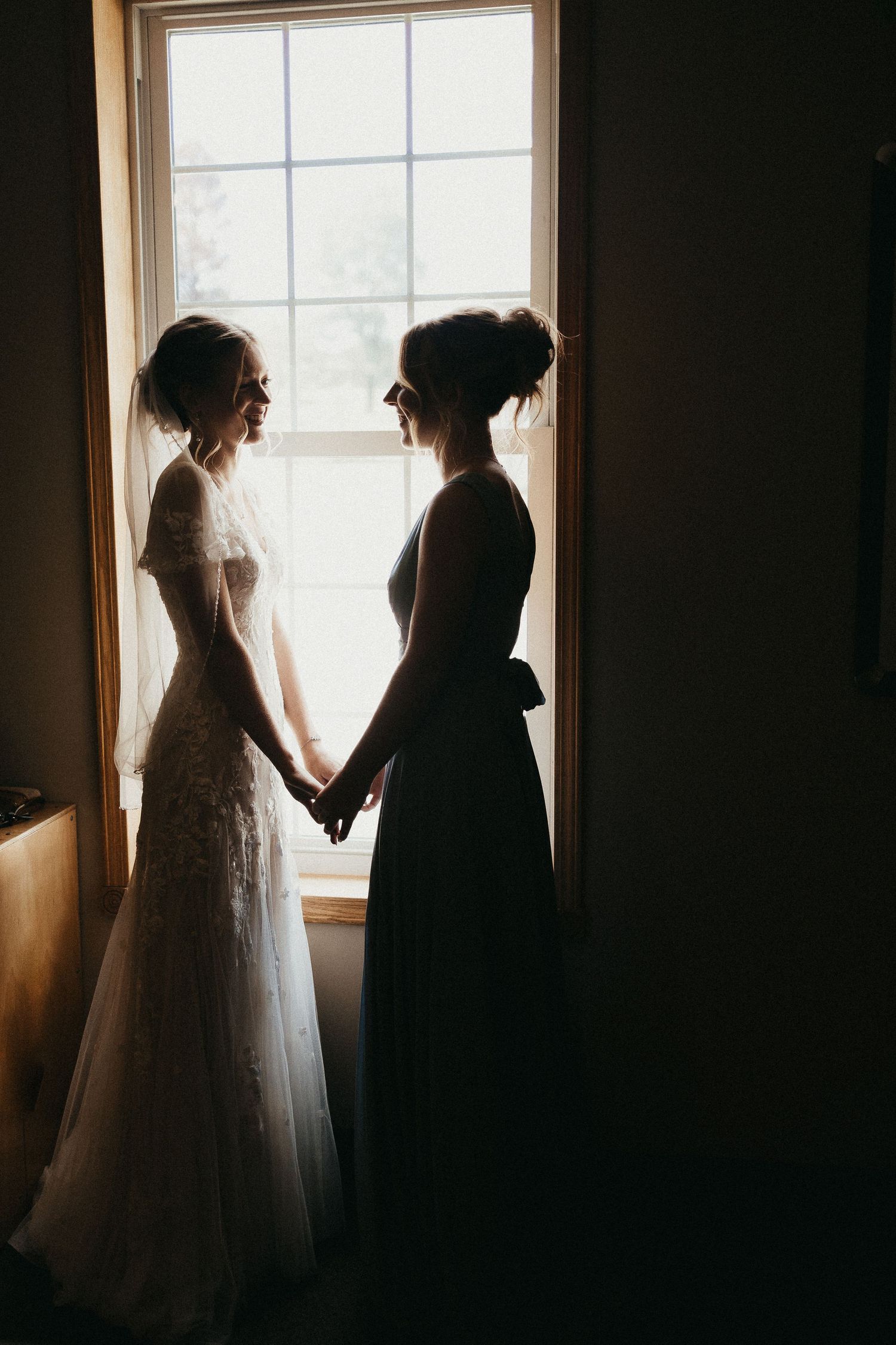 Silhouette of bride and bridesmaid holding hands facing each other in front of sunlit window.