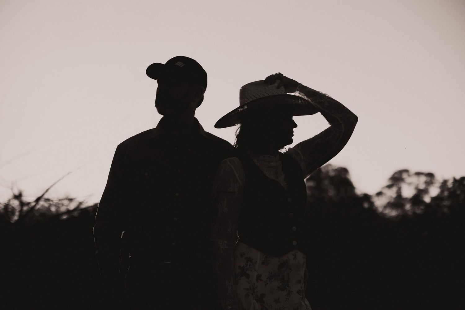 Silhouette of a couple at dusk, one wearing a baseball cap and the other a wide-brim cowboy hat, against a dark sky.