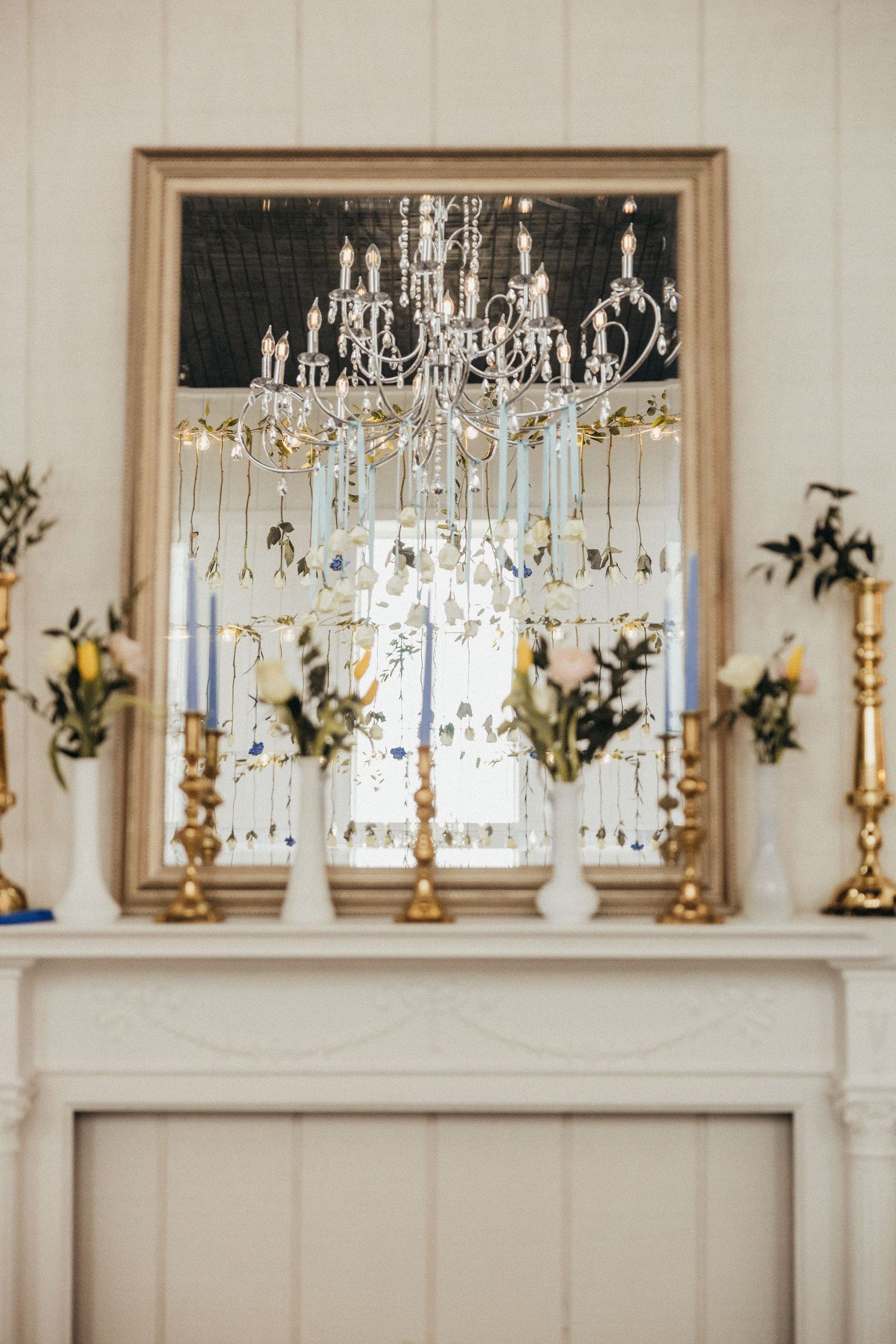 Elegant white fireplace mantel with gold candelabras, floral arrangements, and crystal chandelier reflection in mirror.