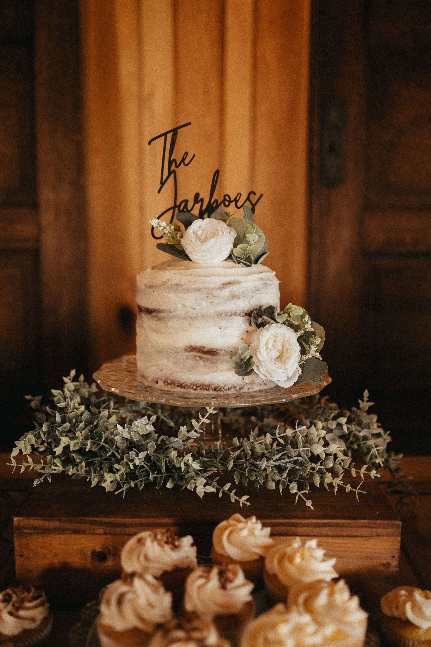 Single-tier semi-naked wedding cake with white roses, greenery, and The Jarboes topper on rustic wood stand.