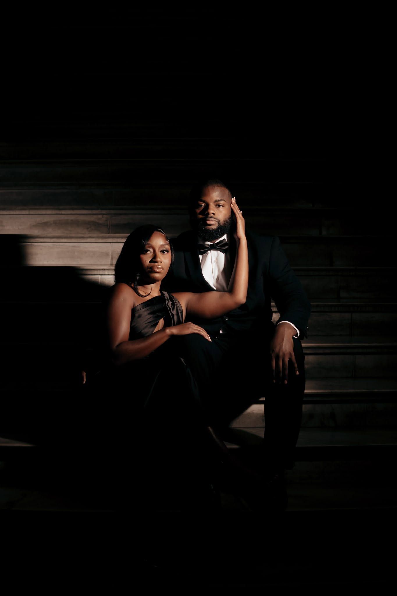 Elegant couple in formal black attire sitting on dark steps, woman in gown leaning on man in tuxedo with bow tie.
