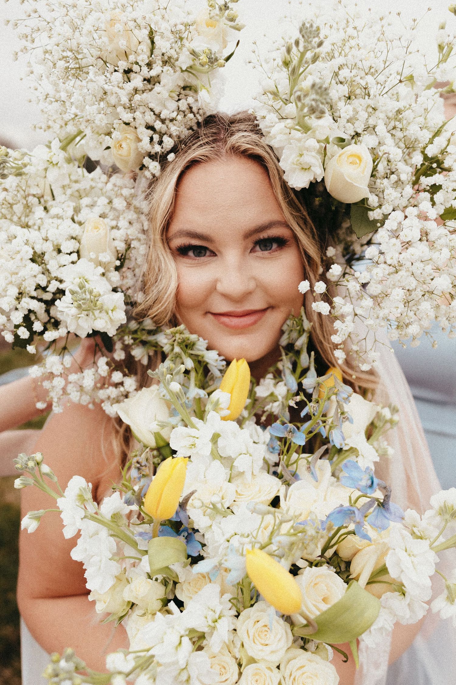 Smiling blonde bride surrounded by large floral crown holding bouquet of white roses and yellow tulips.