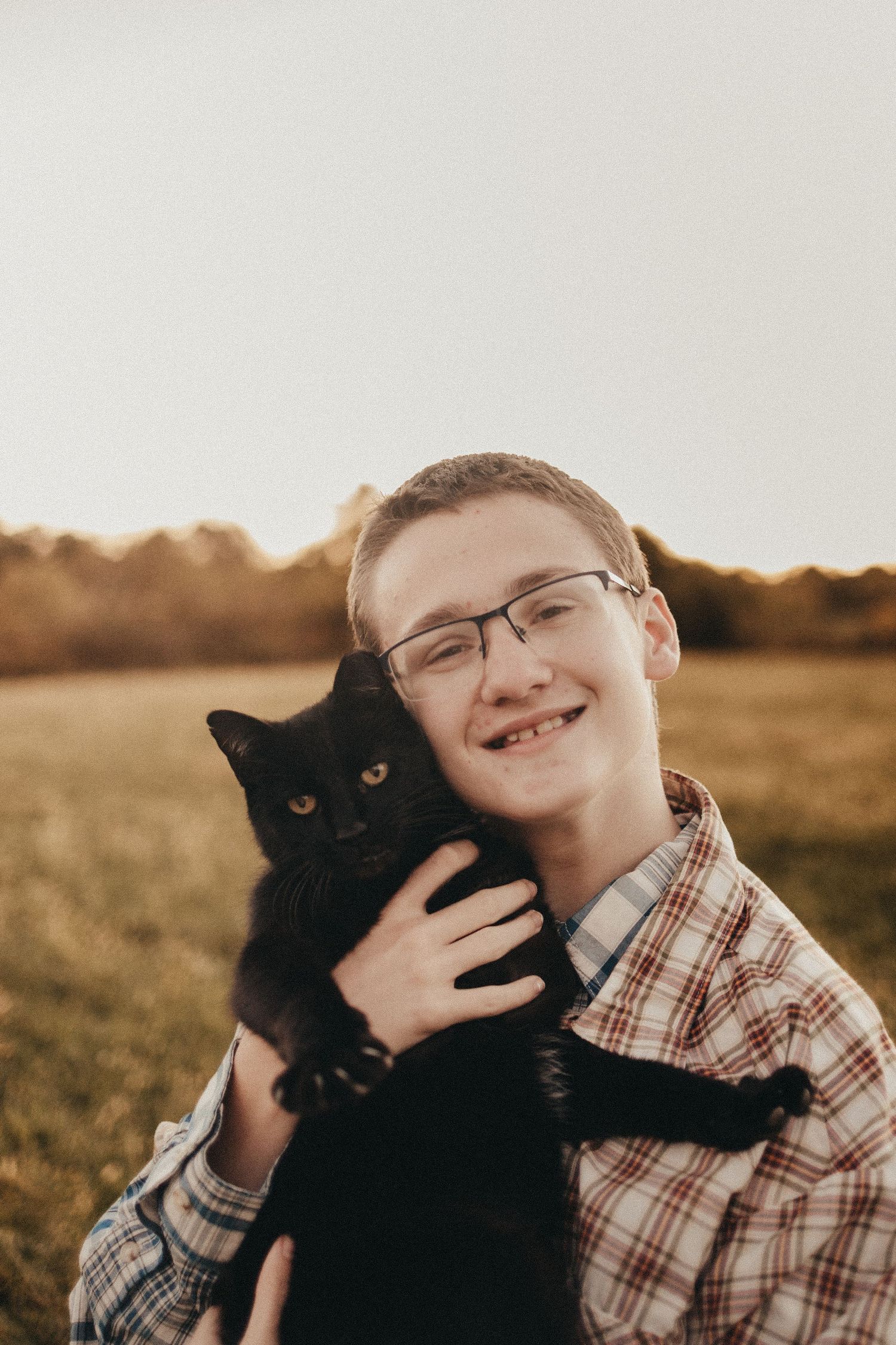 A smiling teenage boy with glasses holds a fluffy black cat during a golden hour outdoor portrait session.