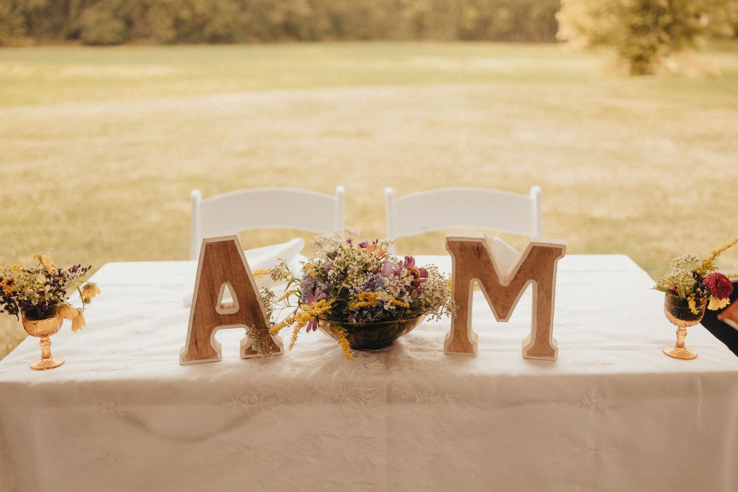 Rustic sweetheart table decorated with letters A and M and wildflower centerpiece at outdoor wedding.