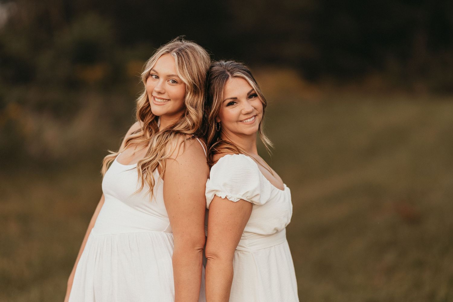 Two smiling women in white dresses standing back to back in a golden outdoor field.