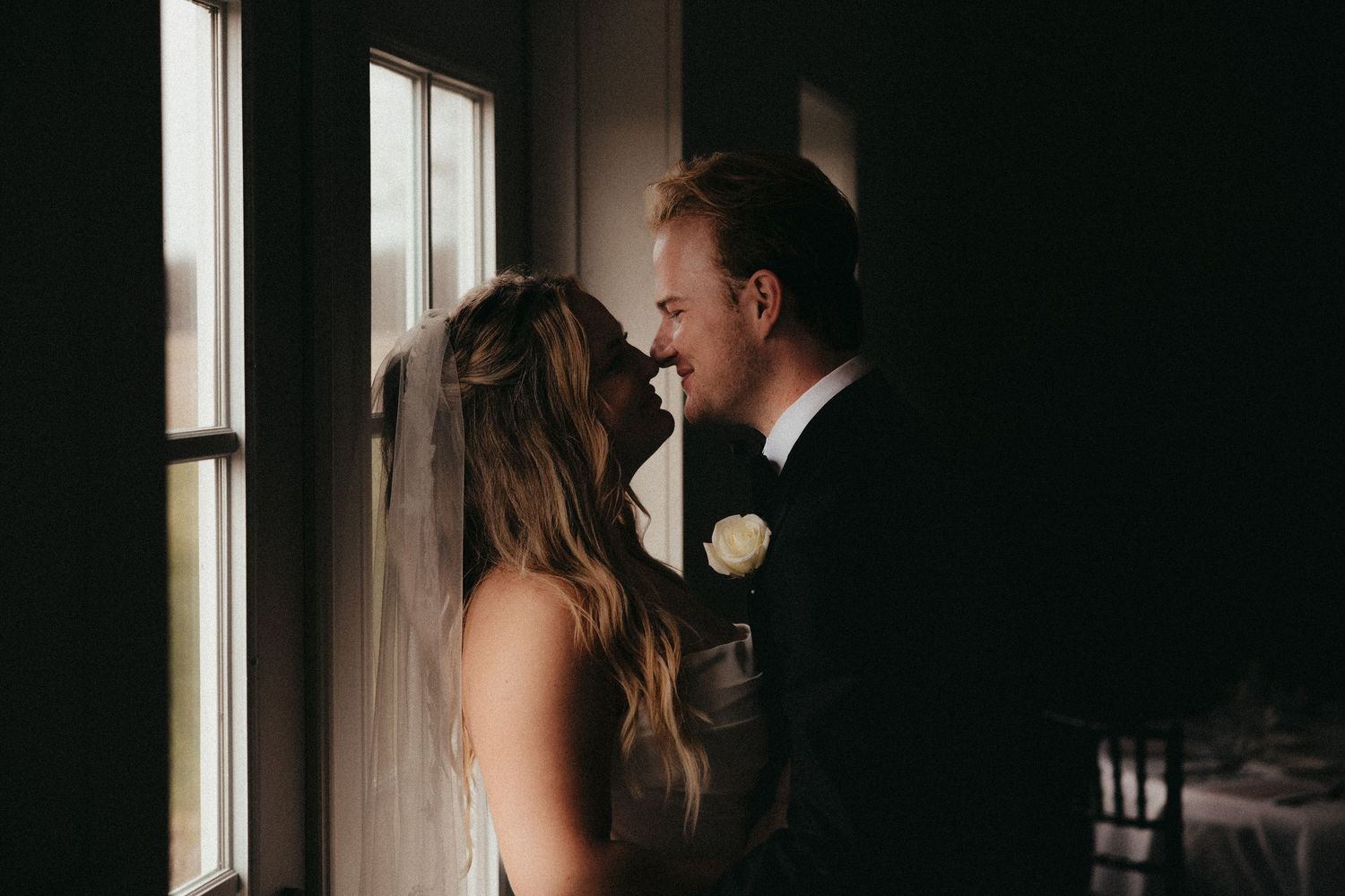 Bride and groom share intimate moment by sunlit window in dark romantic wedding venue.