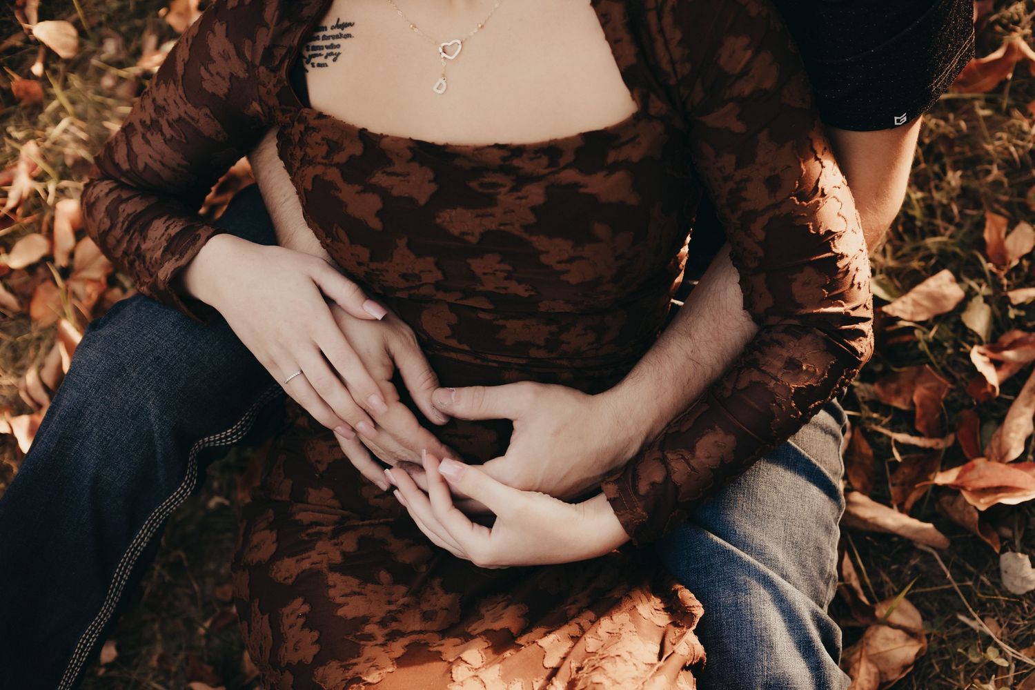 Couple sitting on autumn leaves with hands gently clasped over a pregnant belly, woman in a brown floral lace dress and jeans.