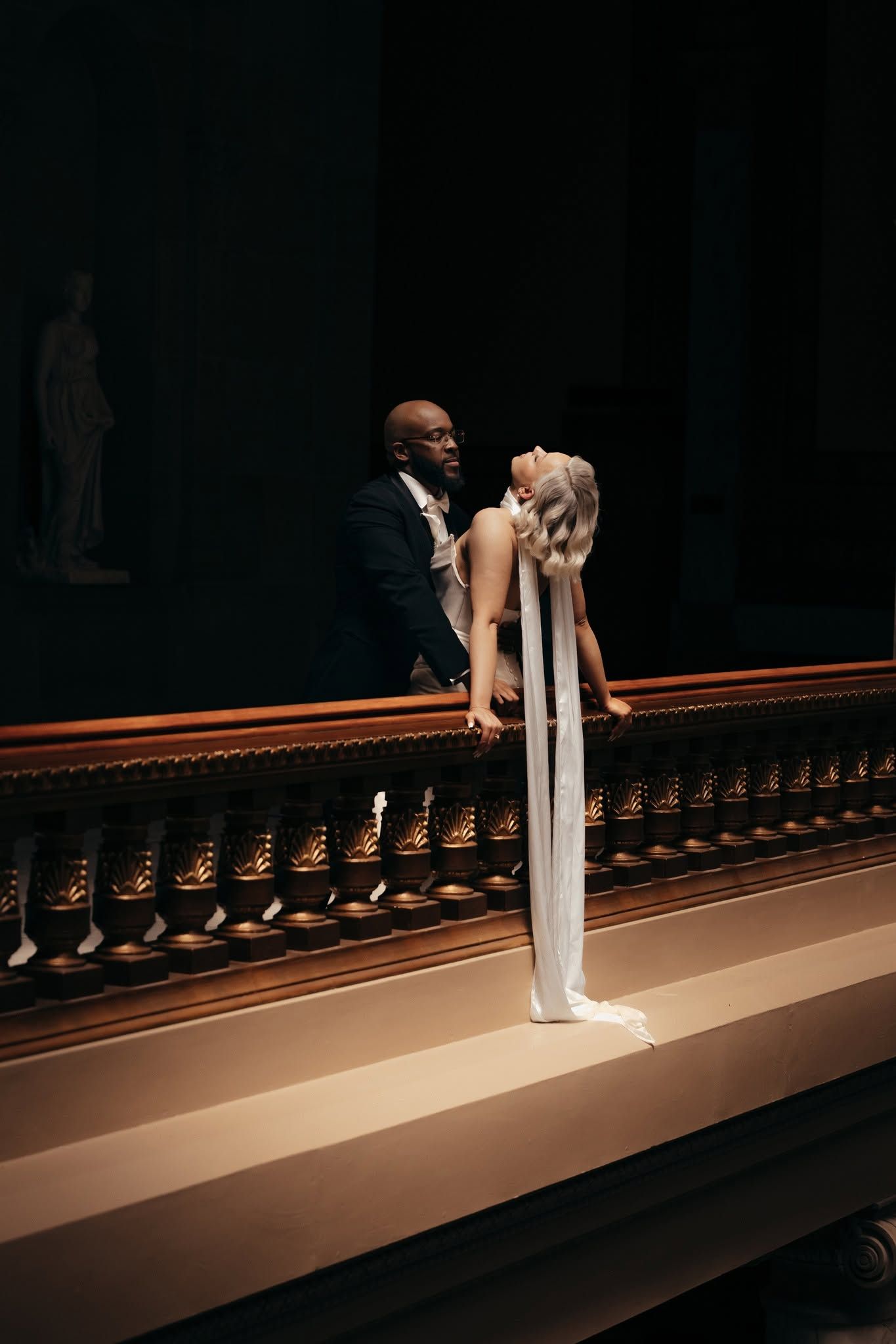 Elegant interracial couple in wedding attire sharing a romantic moment on a grand balcony in a dimly lit venue.