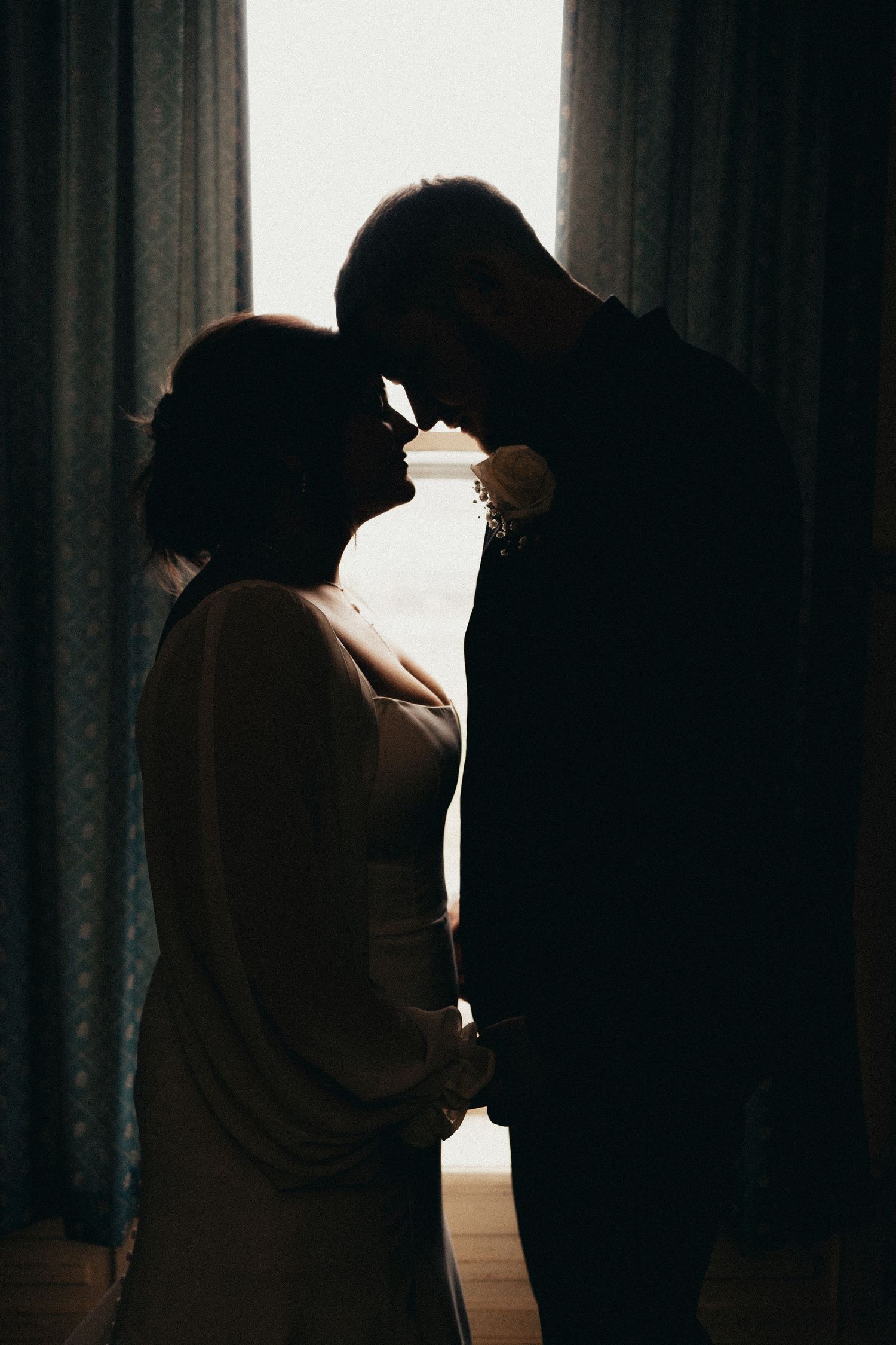 Romantic silhouette of bride and groom touching foreheads in front of a softly lit window.