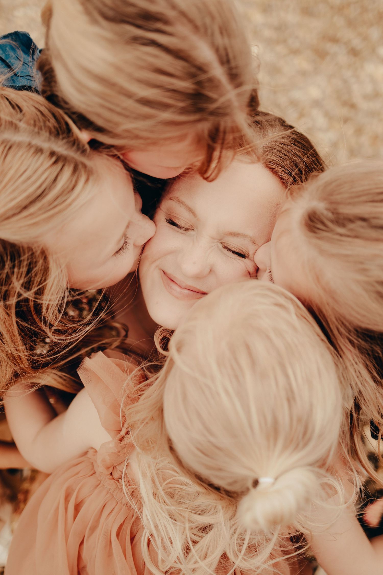 A smiling mother being kissed on both cheeks by her three young daughters in a warm embrace.
