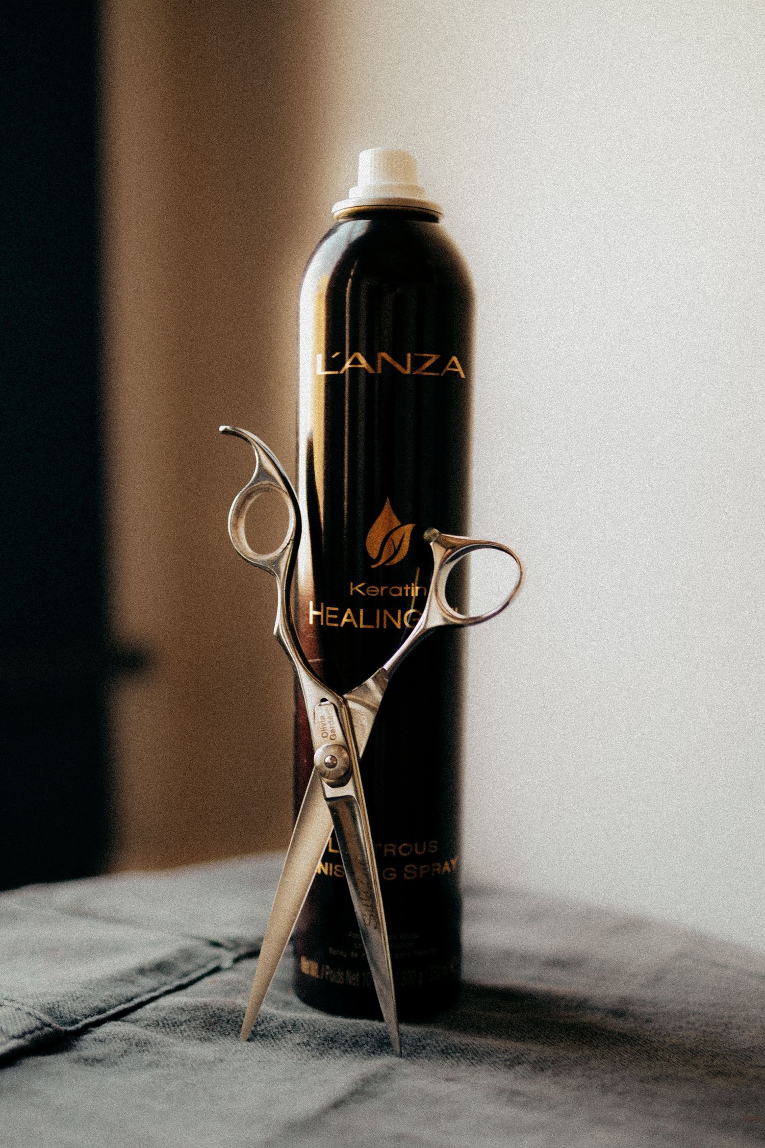 L'Anza Keratin Healing hairspray bottle with silver scissors on a gray surface.