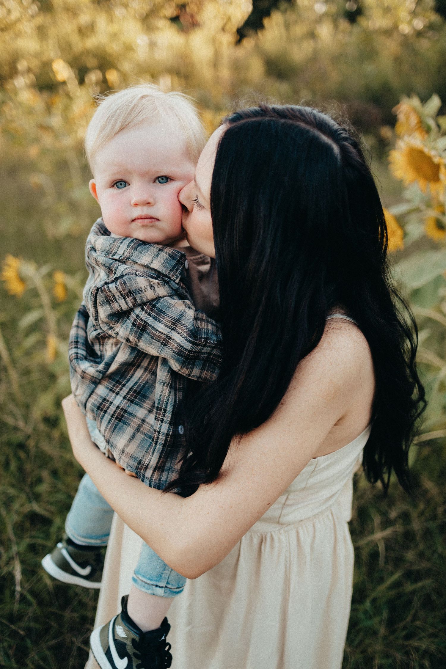 Dark-haired mother kissing her blonde toddler son in a beautiful sunflower field.