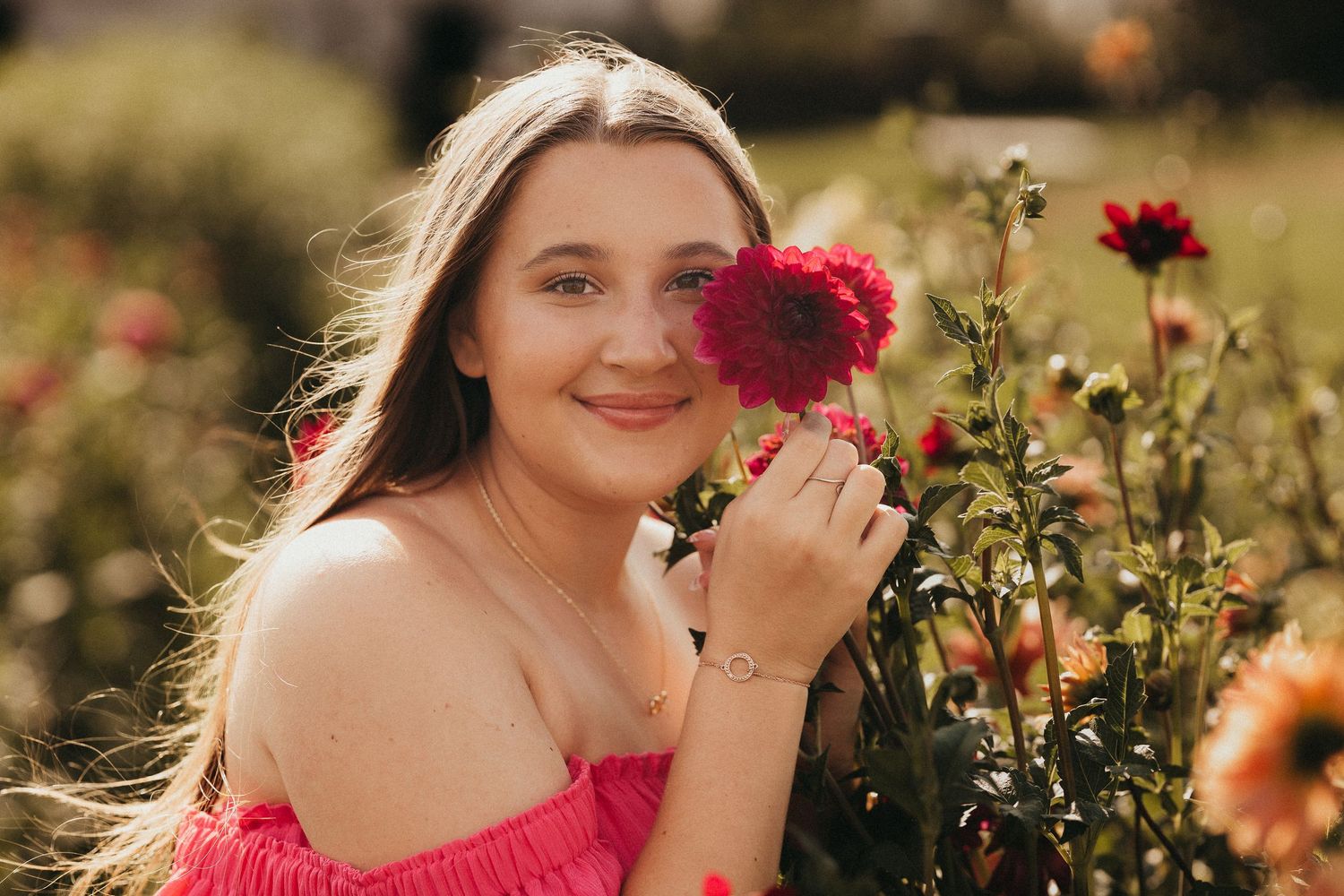 Smiling young woman holding pink dahlia flower in sunny garden for senior portrait.