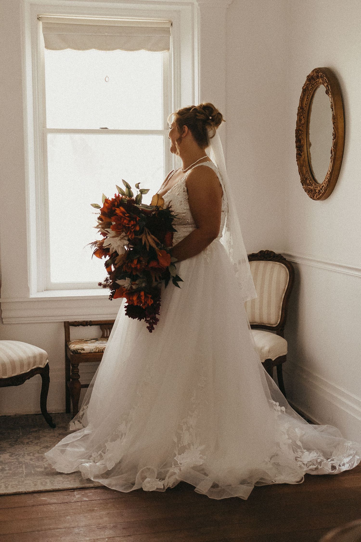 Bride in a white lace ball gown with a long train holds an autumn bouquet of red and orange flowers by a window.