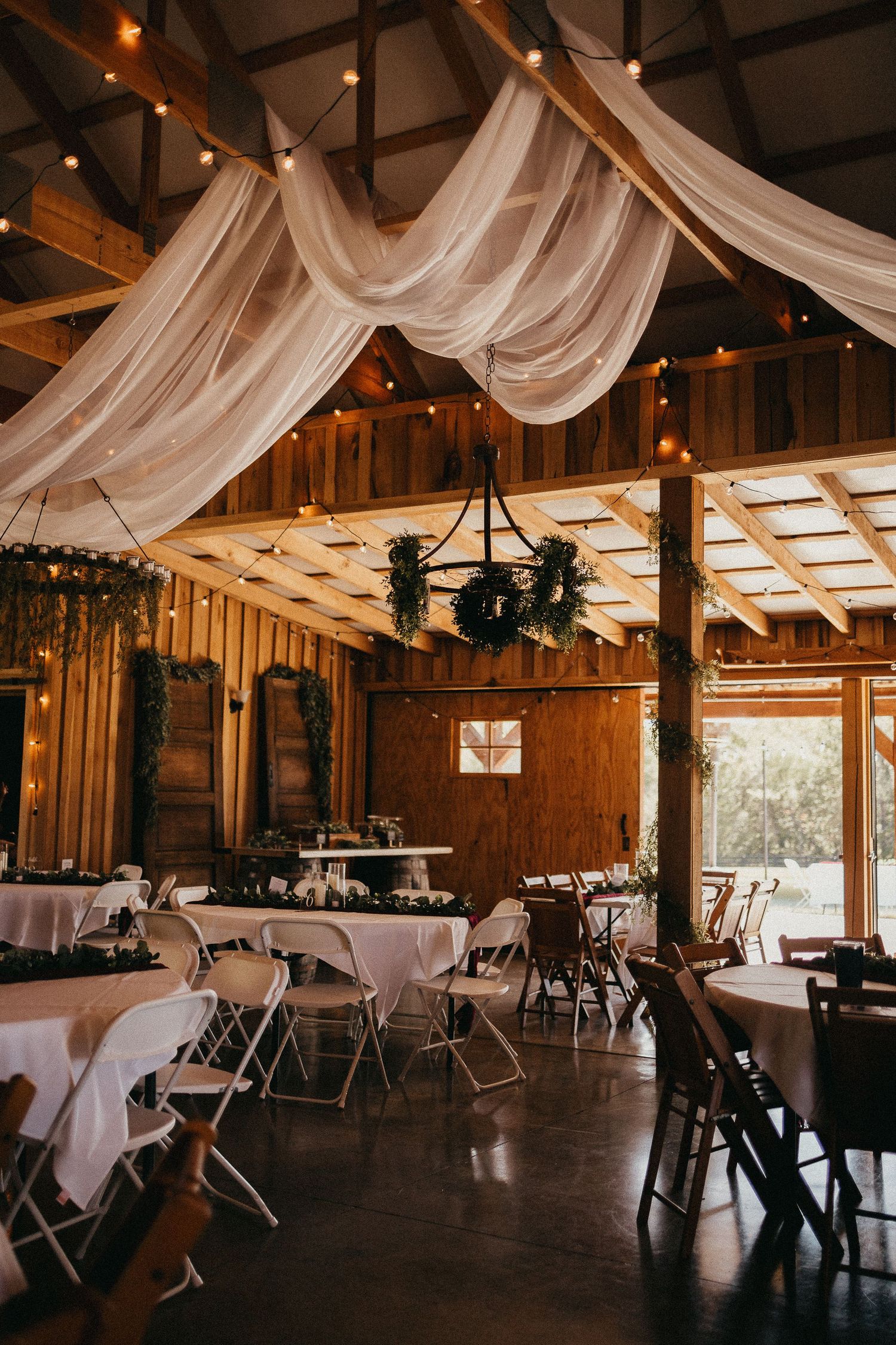 Rustic barn wedding reception venue with white draped ceiling, string lights, round tables, and wooden chandelier.