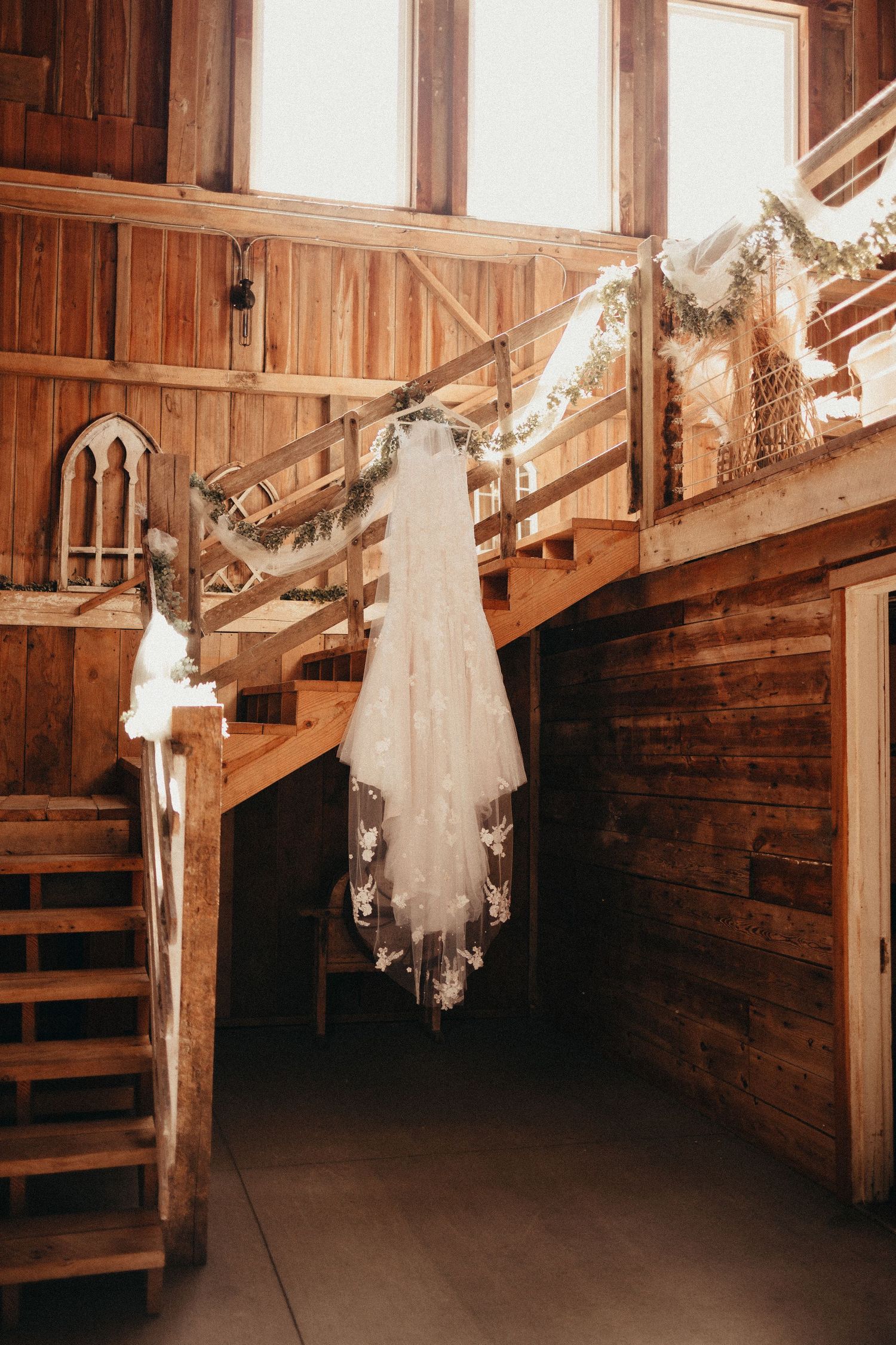 Elegant lace wedding dress and veil hanging from wooden staircase inside a rustic barn wedding venue.