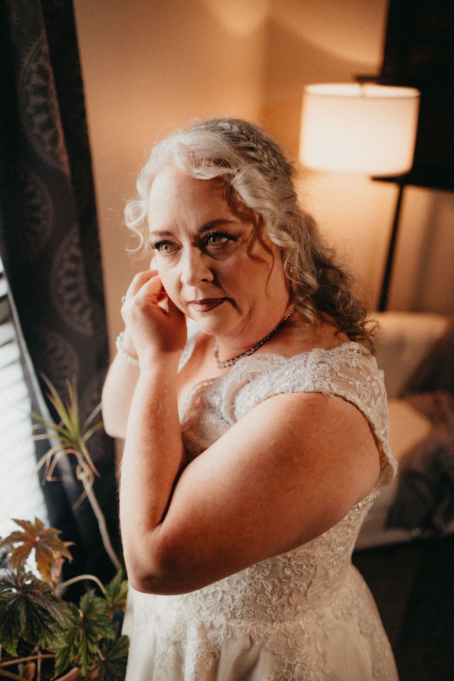 Beautiful mature bride in lace cap sleeve wedding dress adjusting earring in warm lit room.