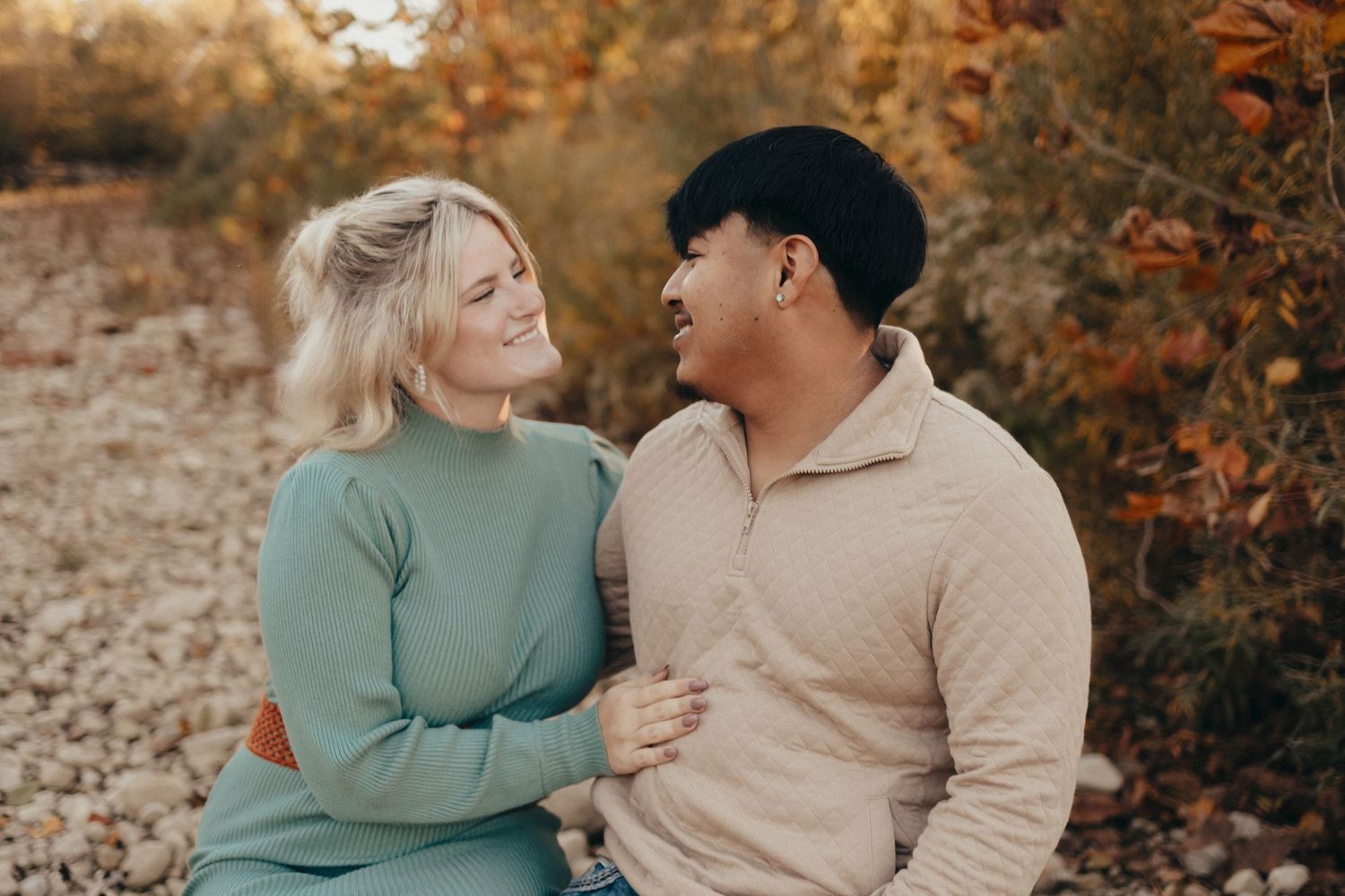 A happy couple sits together smiling at each other surrounded by colorful autumn foliage in a warm outdoor setting.
