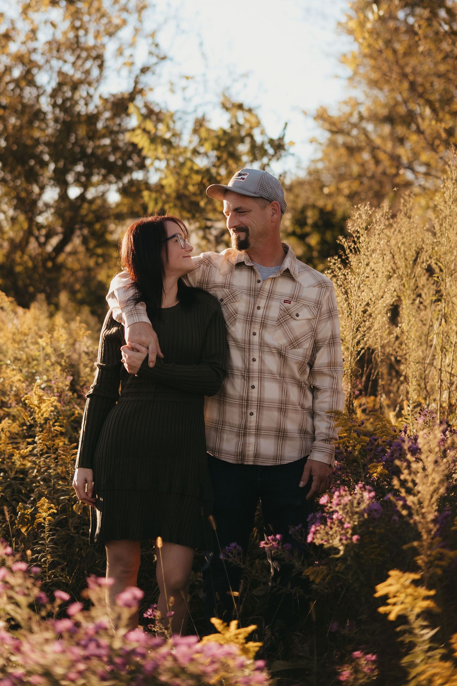 Couple embracing in a sunlit wildflower field during golden hour, woman in black dress and man in plaid shirt smiling at each other.