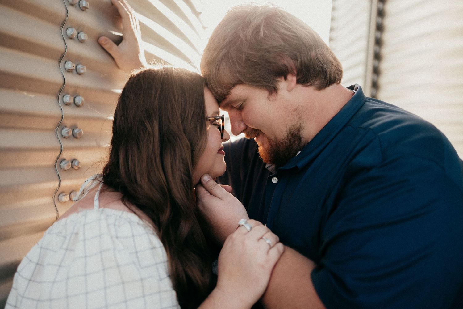 Couple sharing a tender forehead-to-forehead moment outdoors near rustic metal grain bins.