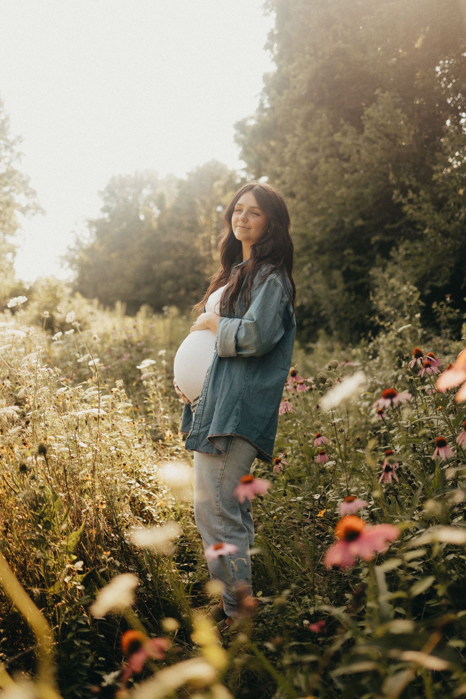 Smiling pregnant woman in denim jacket standing in a sunlit wildflower field.