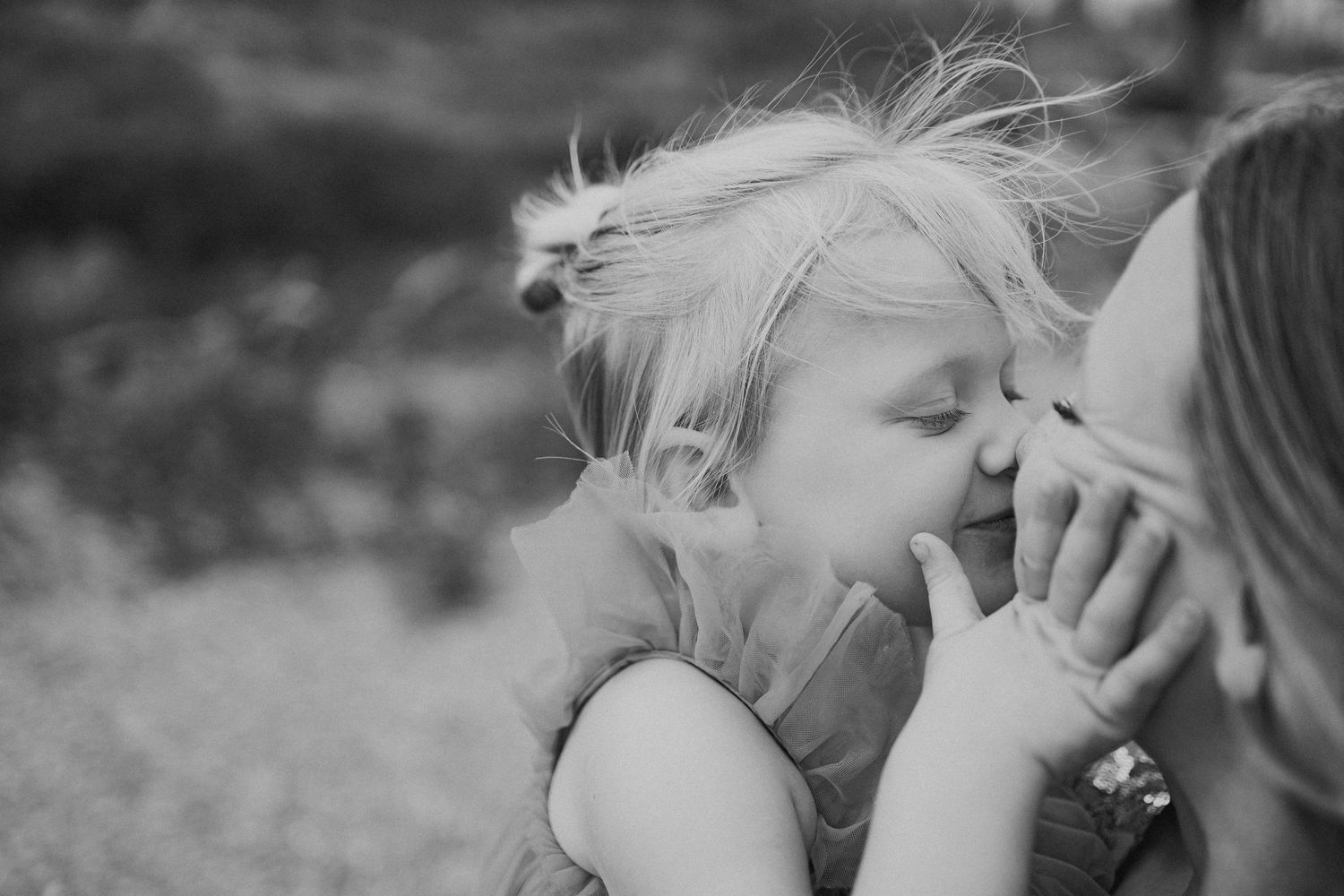 Black and white photo of a young blonde child kissing a womans cheek outdoors.