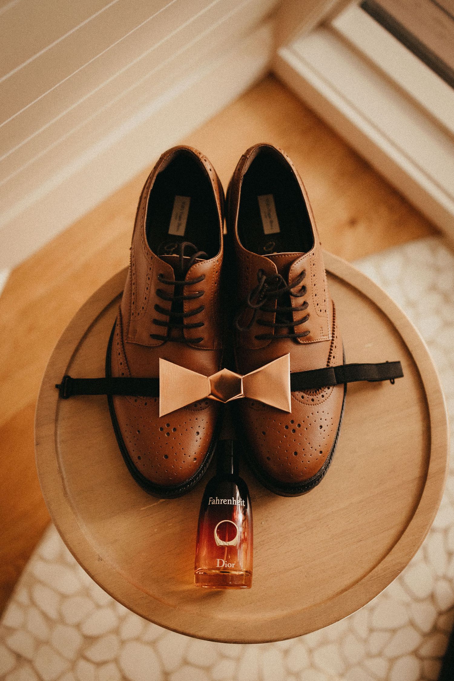 Brown leather brogue groom shoes with nude bow tie and Dior Fahrenheit cologne on wooden tray.