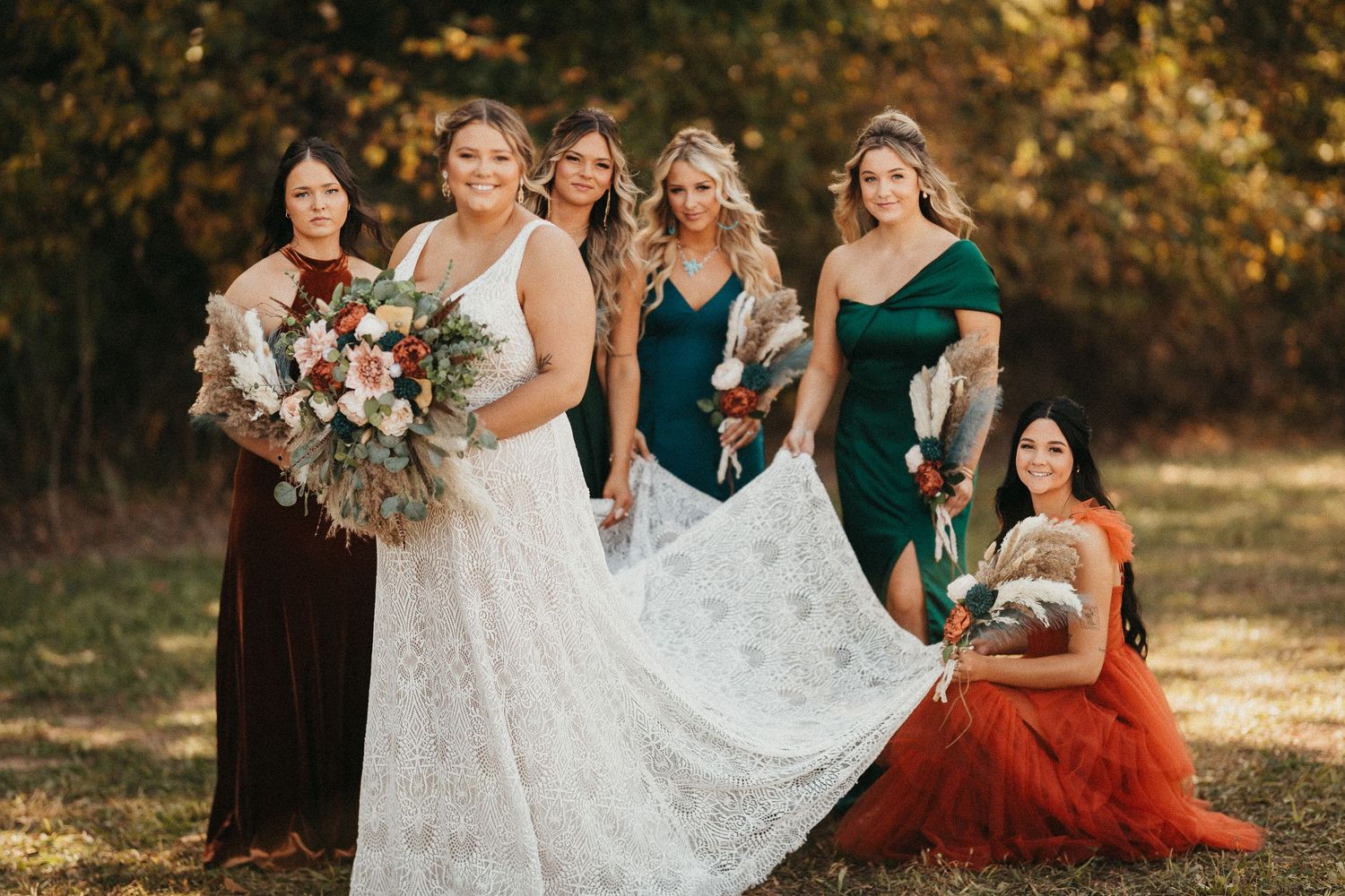 Bride in white lace gown poses with five bridesmaids in jewel-toned dresses outdoors in autumn.