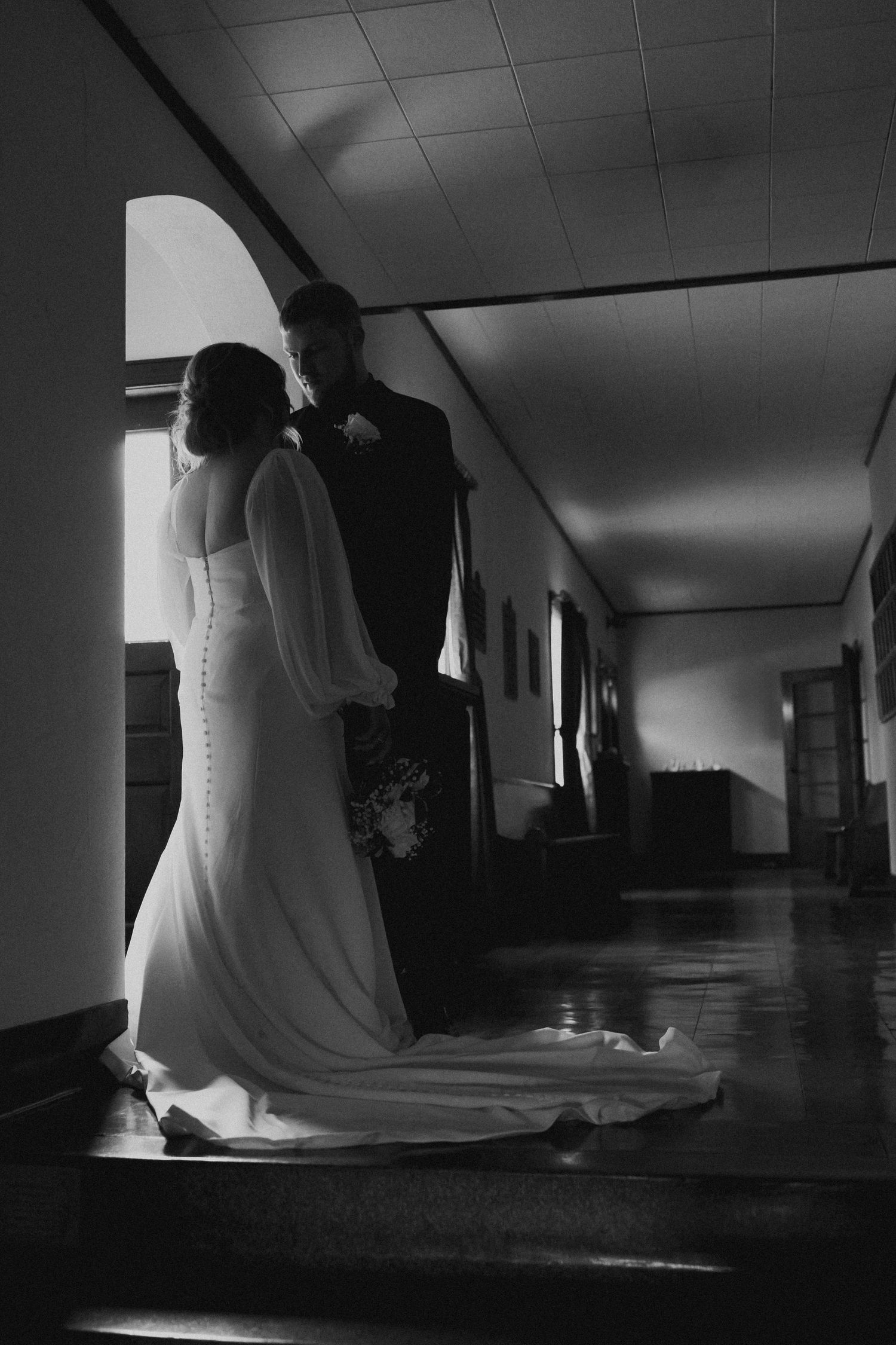Black and white photo of bride and groom standing together in sunlit church hallway.
